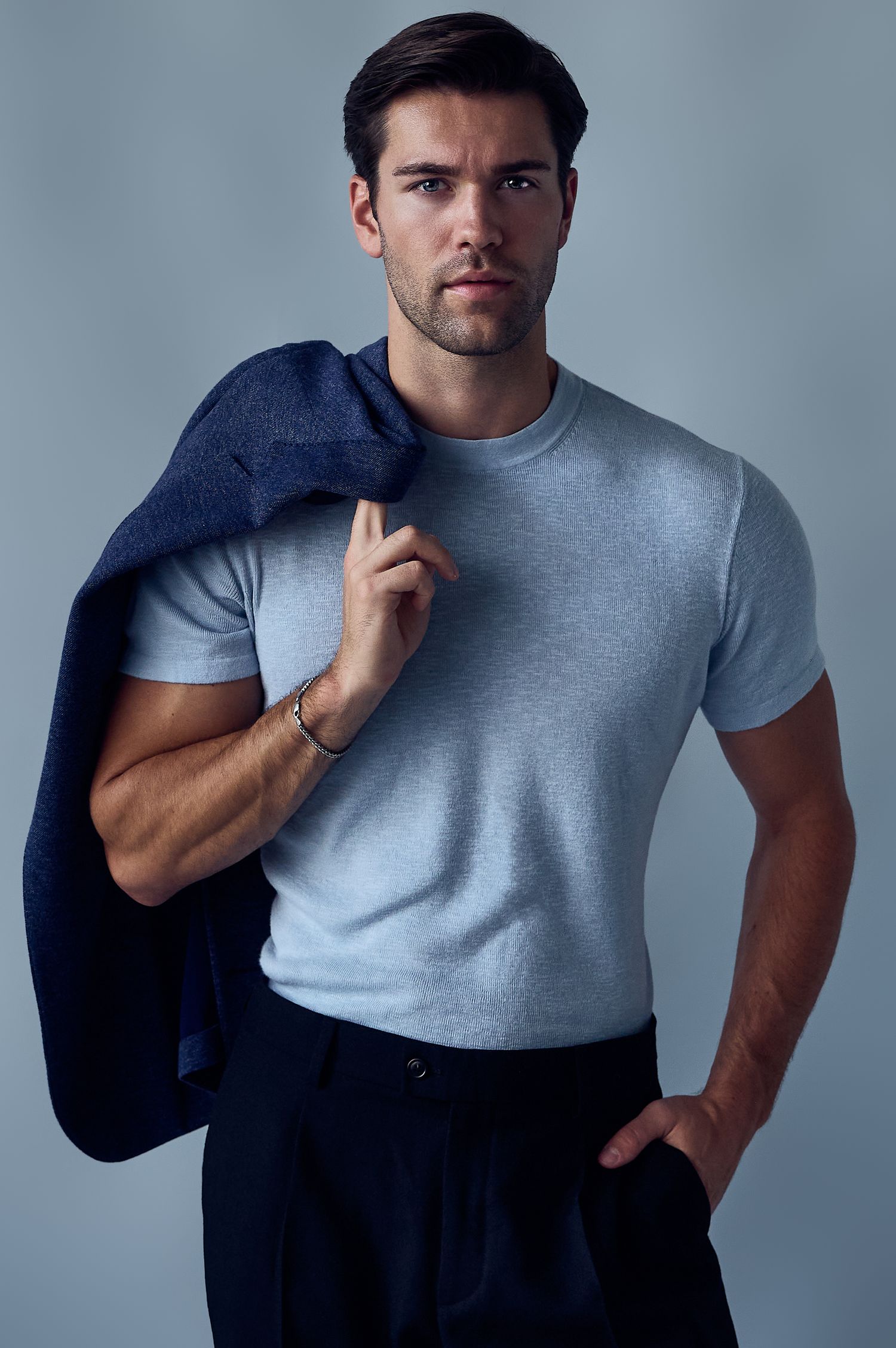 Confident young man in gray t-shirt holding navy blazer over shoulder, posing against gray background.