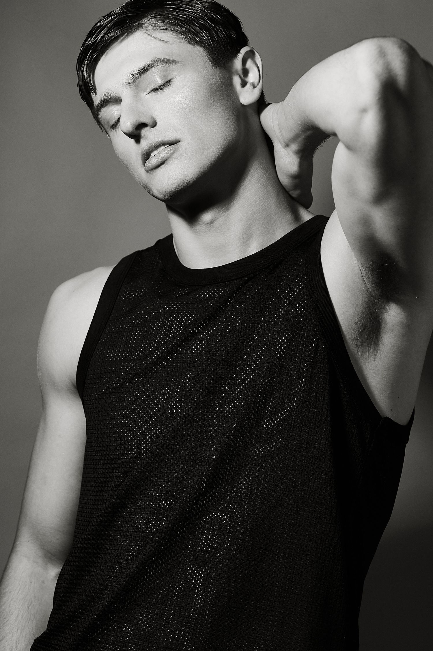 Black and white photo of athletic male model in mesh tank top with eyes closed, flexing arm.