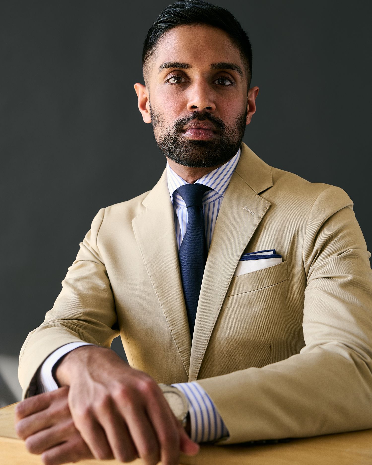 Bearded man in tan blazer and blue tie seated at table against gray background.