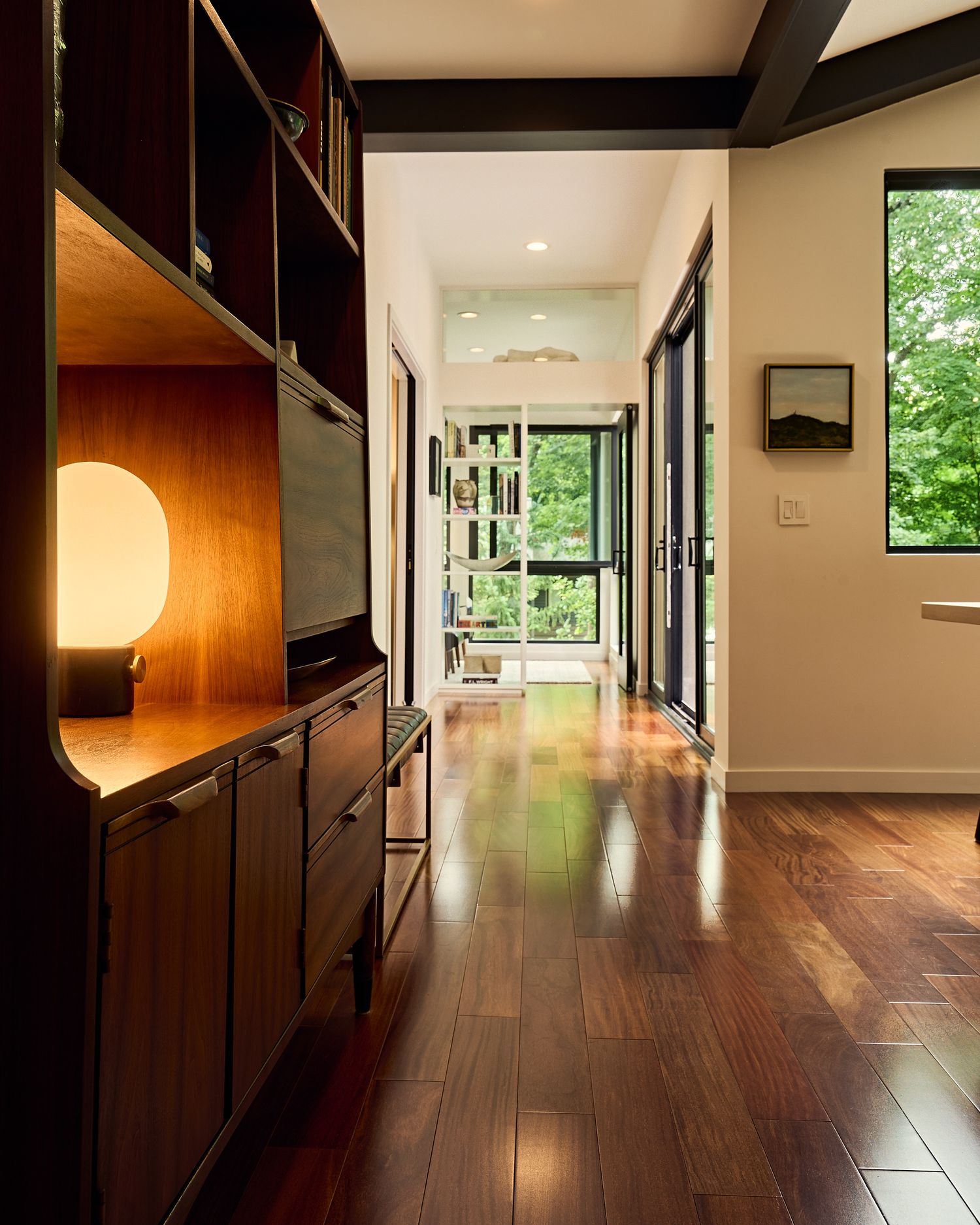 Modern hallway with rich hardwood floors, wood built-in shelving, and floor-to-ceiling windows overlooking lush greenery.