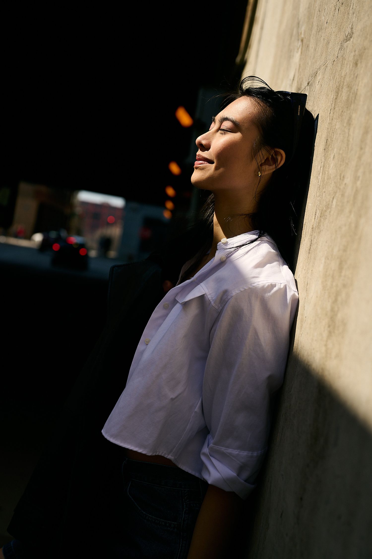 A young Asian woman in a white shirt leans against a sunlit wall with eyes closed, bathed in warm natural light.