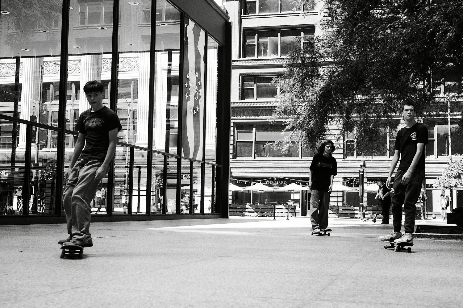 Black and white photo of three skateboarders riding in front of a modern glass building.