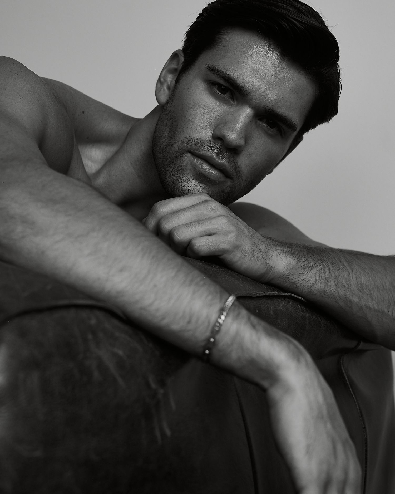 Shirtless muscular man with dark hair poses thoughtfully on leather chair in black and white portrait photo.