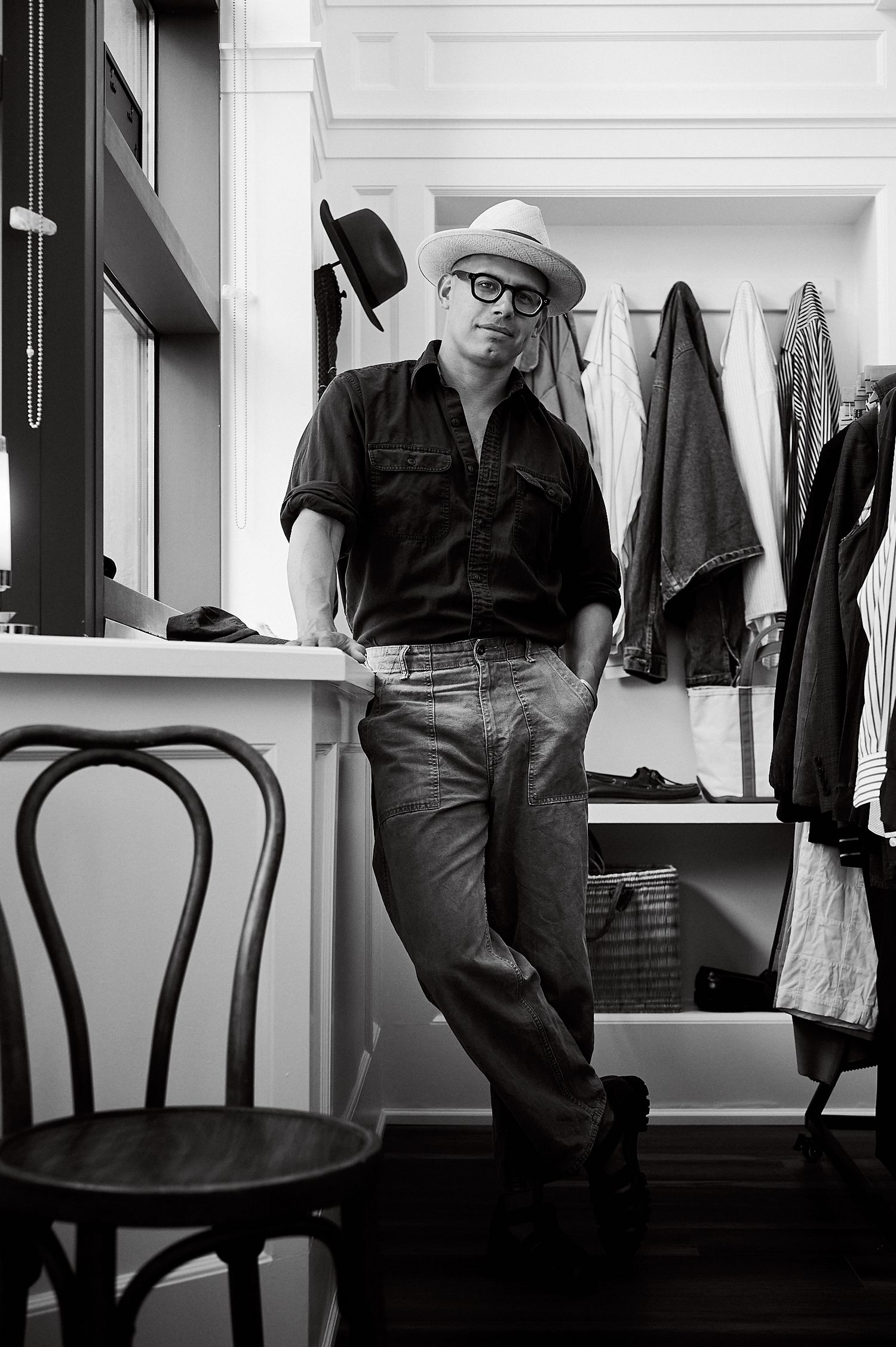 Black and white photo of a man in a hat and glasses leaning against a wall in a clothing store with coats hanging behind him.