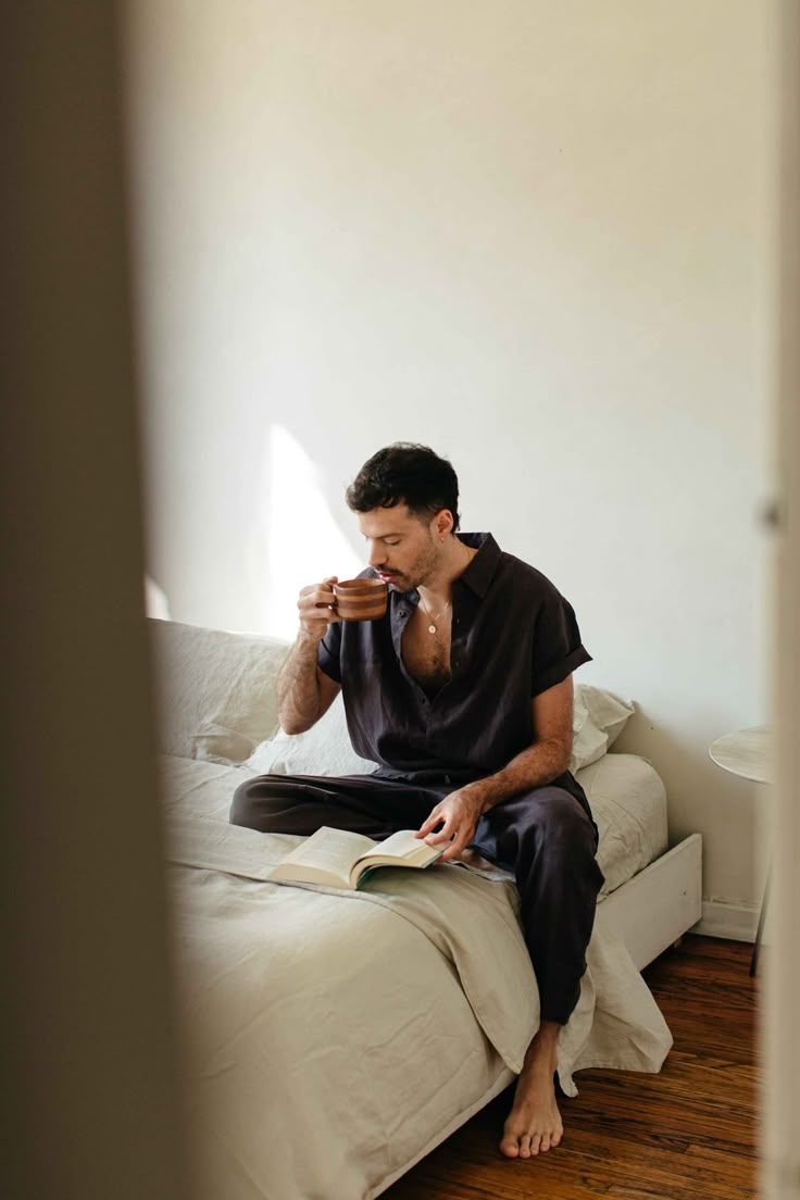 Man in black pajamas sitting on bed, sipping coffee and reading a book in a sunlit bedroom.