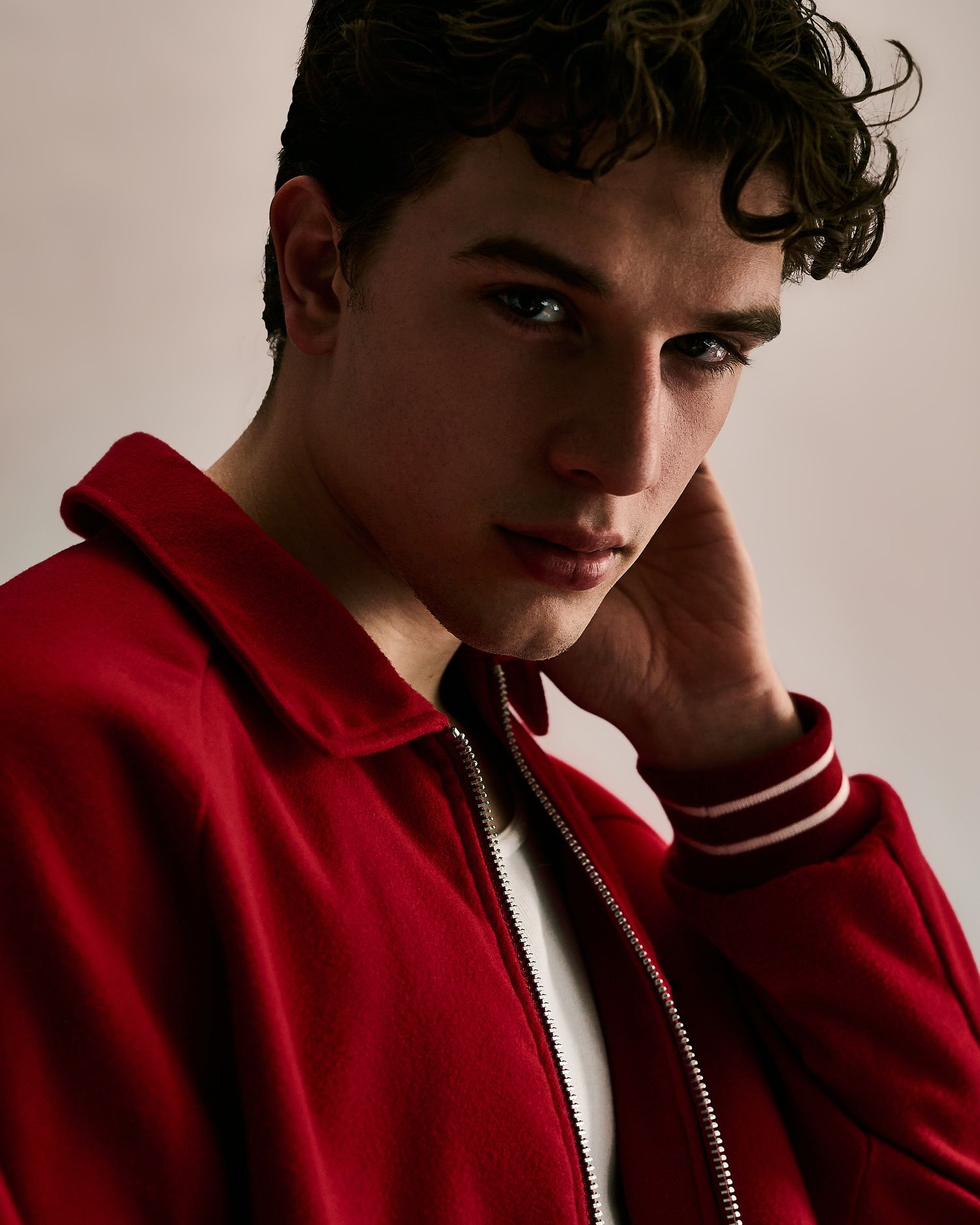 Young man with curly dark hair wearing a red zip-up jacket, posing with hand near face.