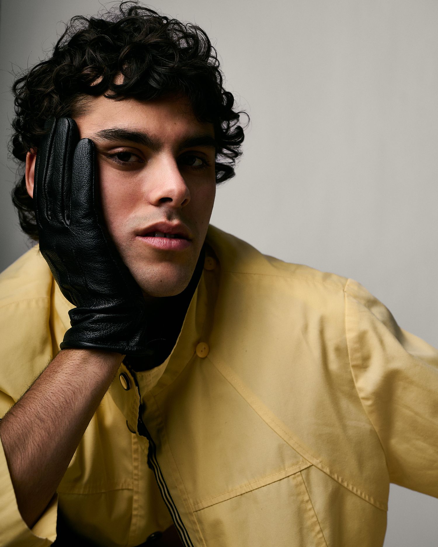 Young man with curly hair wearing a yellow jacket and black leather glove touching his face.