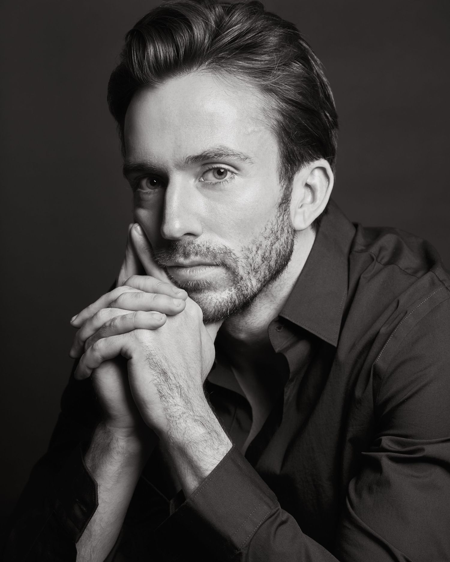 Black and white portrait of a bearded man in a dark shirt, hands clasped under chin, studio photography.