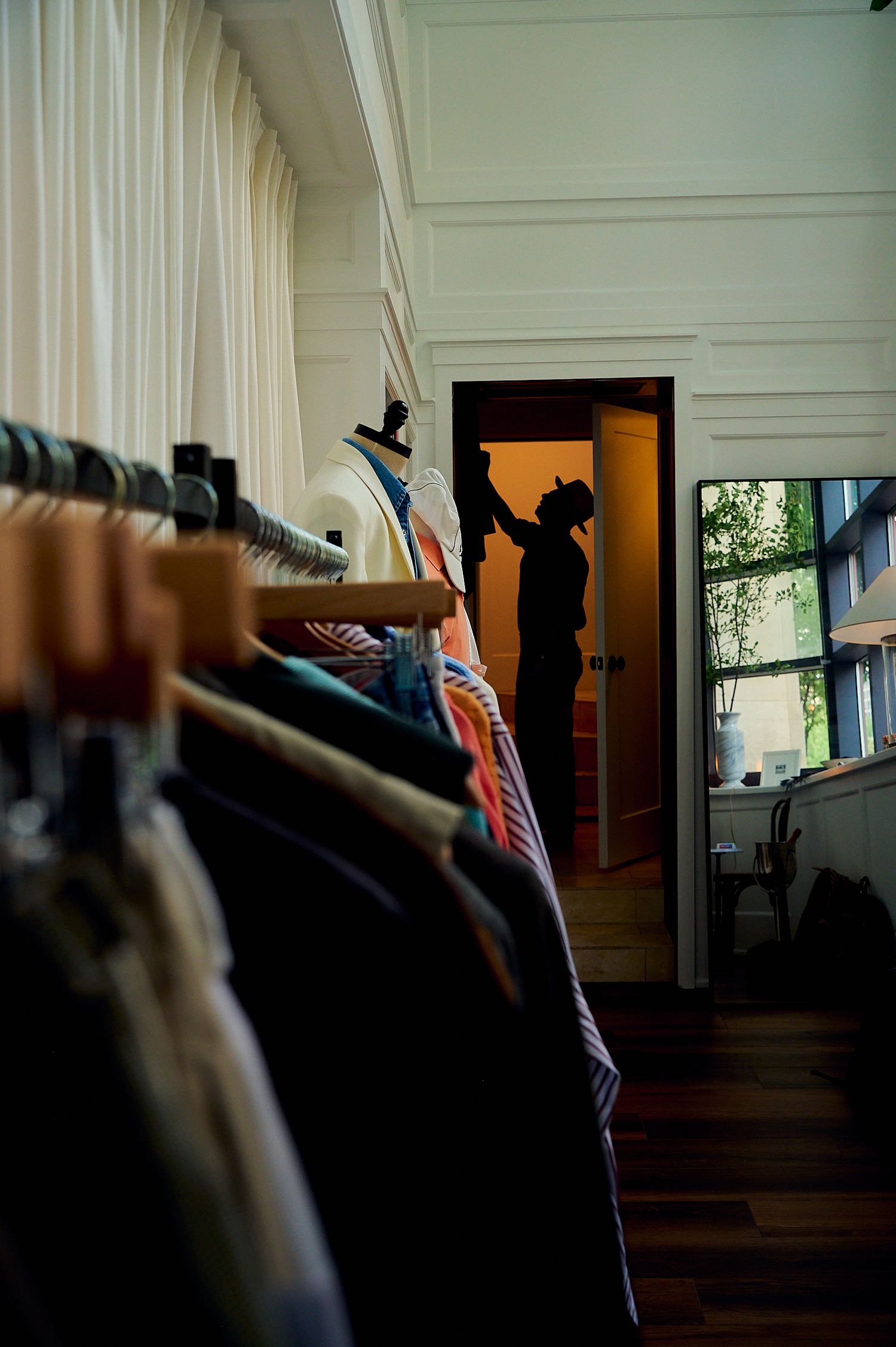 Silhouette of a person browsing clothes on wooden hangers in a dimly lit boutique clothing store.