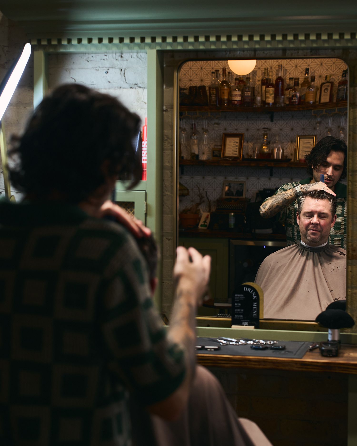 Barber styling a clients hair reflected in a vintage mirror with shelved products behind.