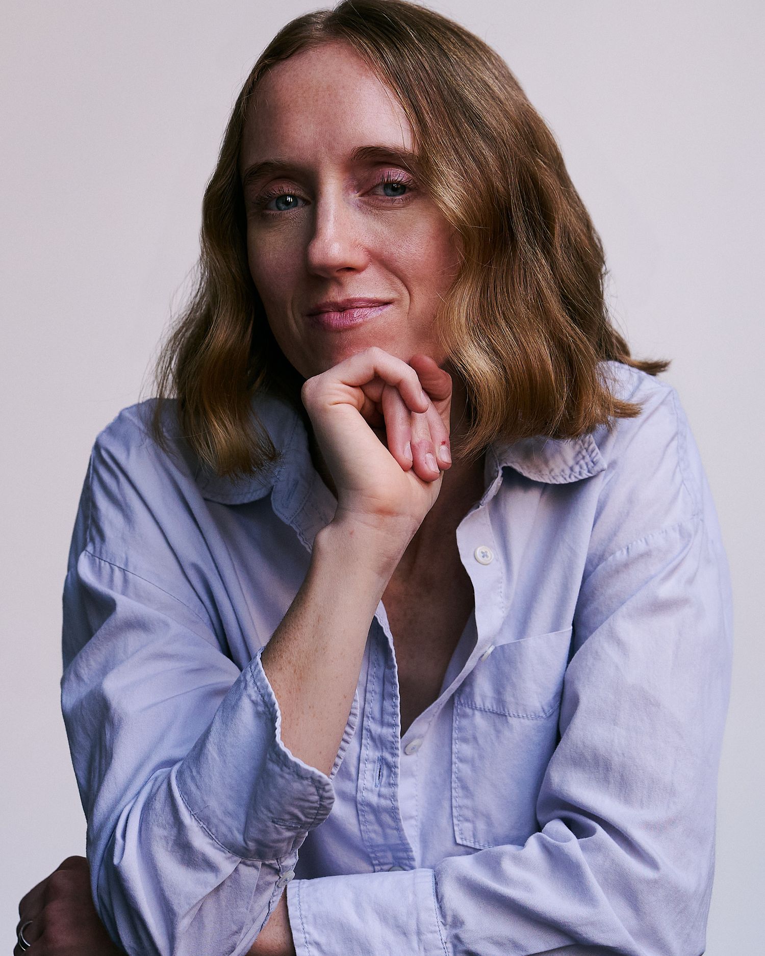 A woman with shoulder-length hair poses thoughtfully with hand on chin, wearing a light blue button-up shirt.