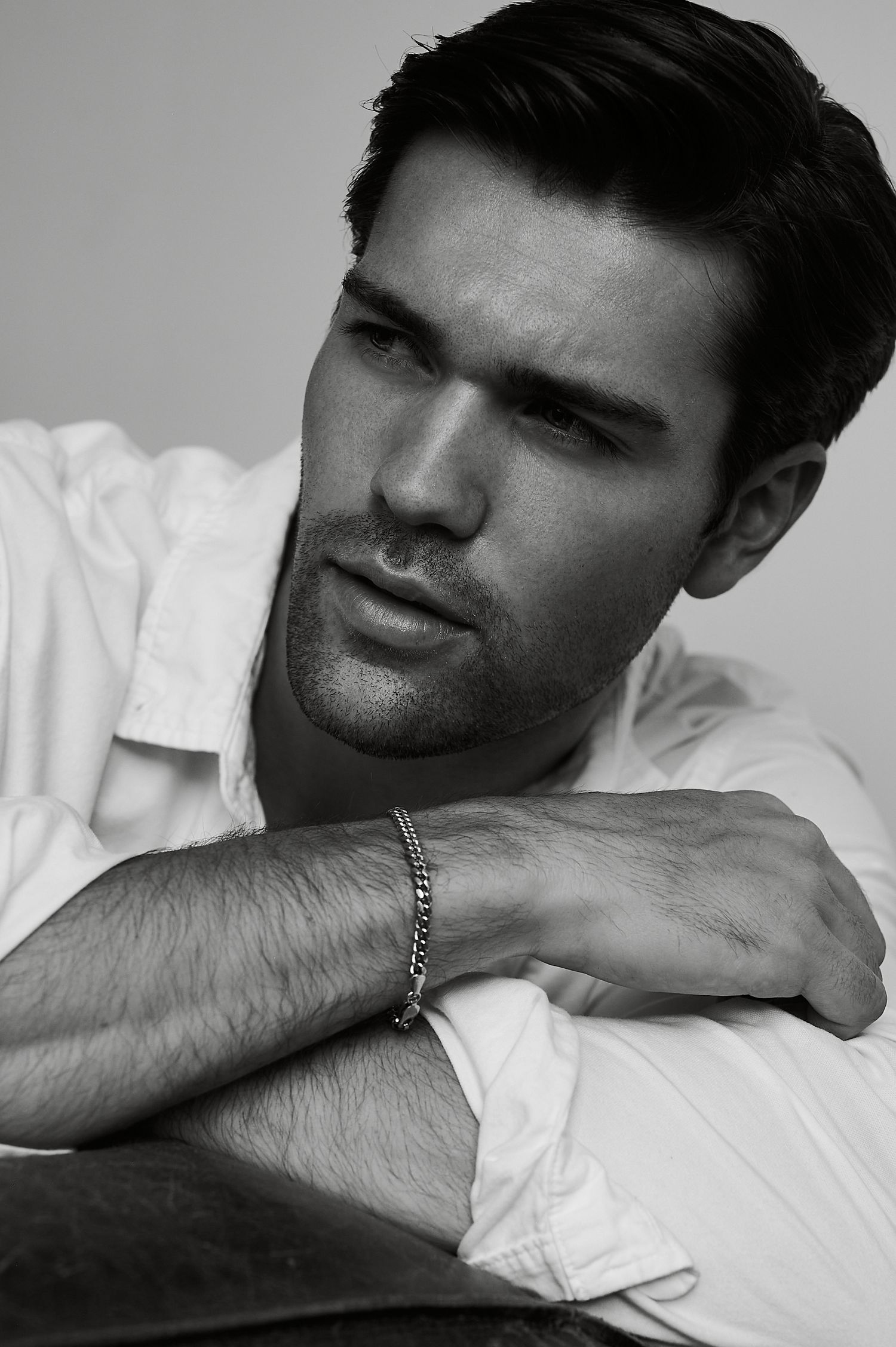 Black and white portrait of a handsome man in white shirt with chain bracelet, arms crossed, looking away.