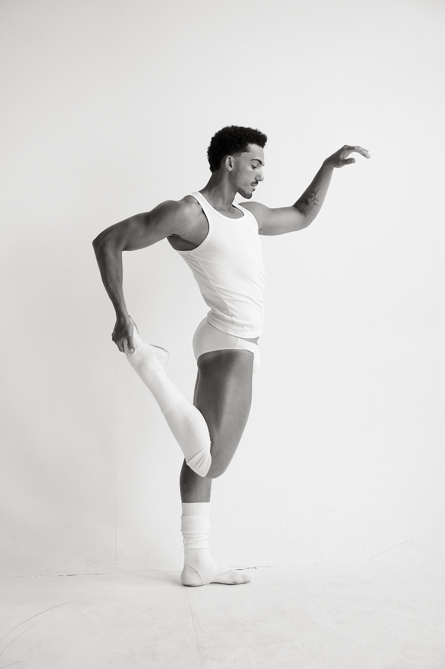 Black and white studio photo of a male ballet dancer in white athletic wear performing a pose.