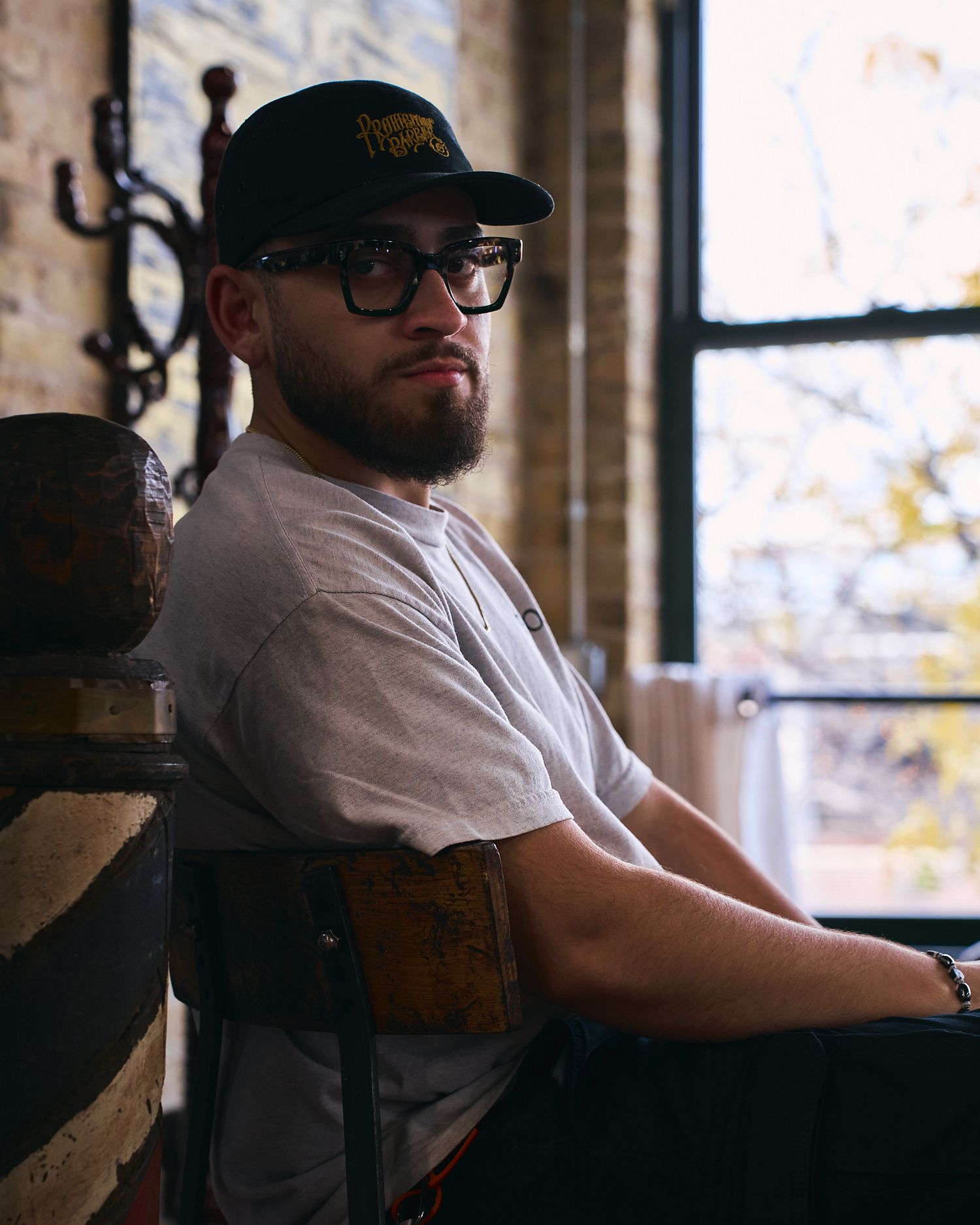 Bearded man wearing glasses and a cap sitting in a vintage barber chair by a window.