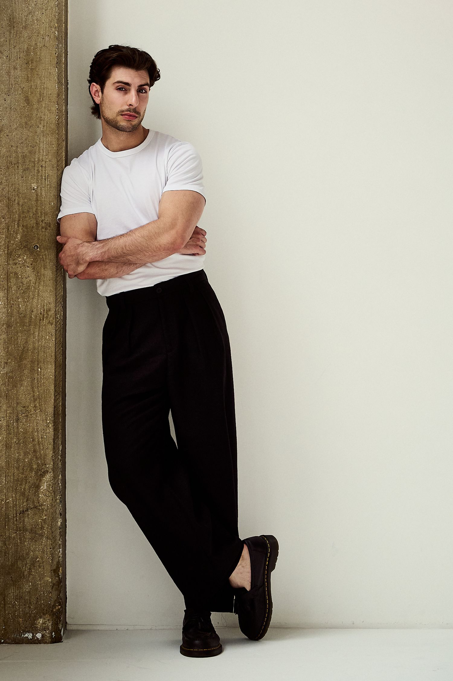 Young man in white t-shirt and black wide-leg trousers leaning against wall with arms crossed.