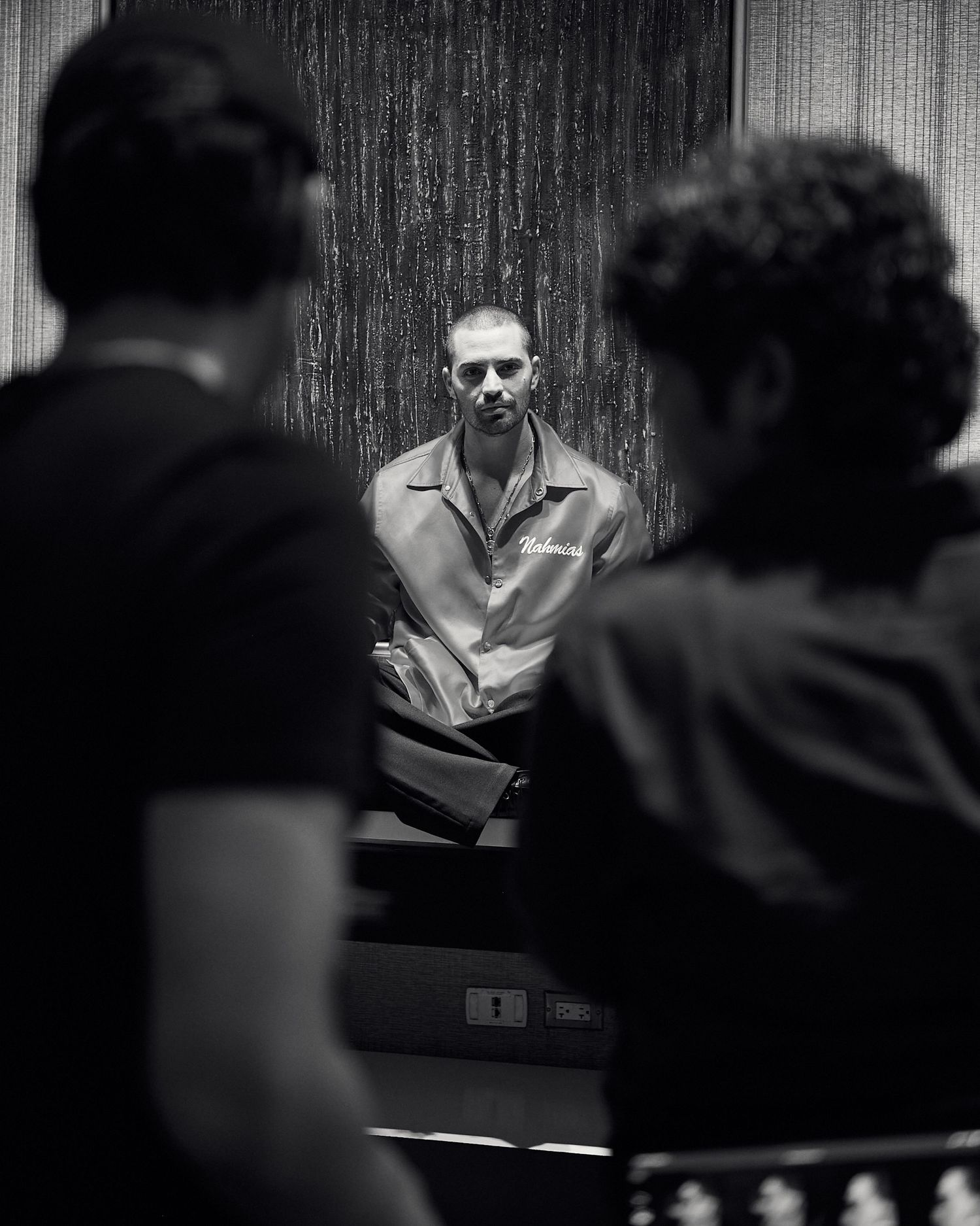 Black and white photo of a man in a button-up shirt facing two blurred figures in a dramatic, moody indoor setting.