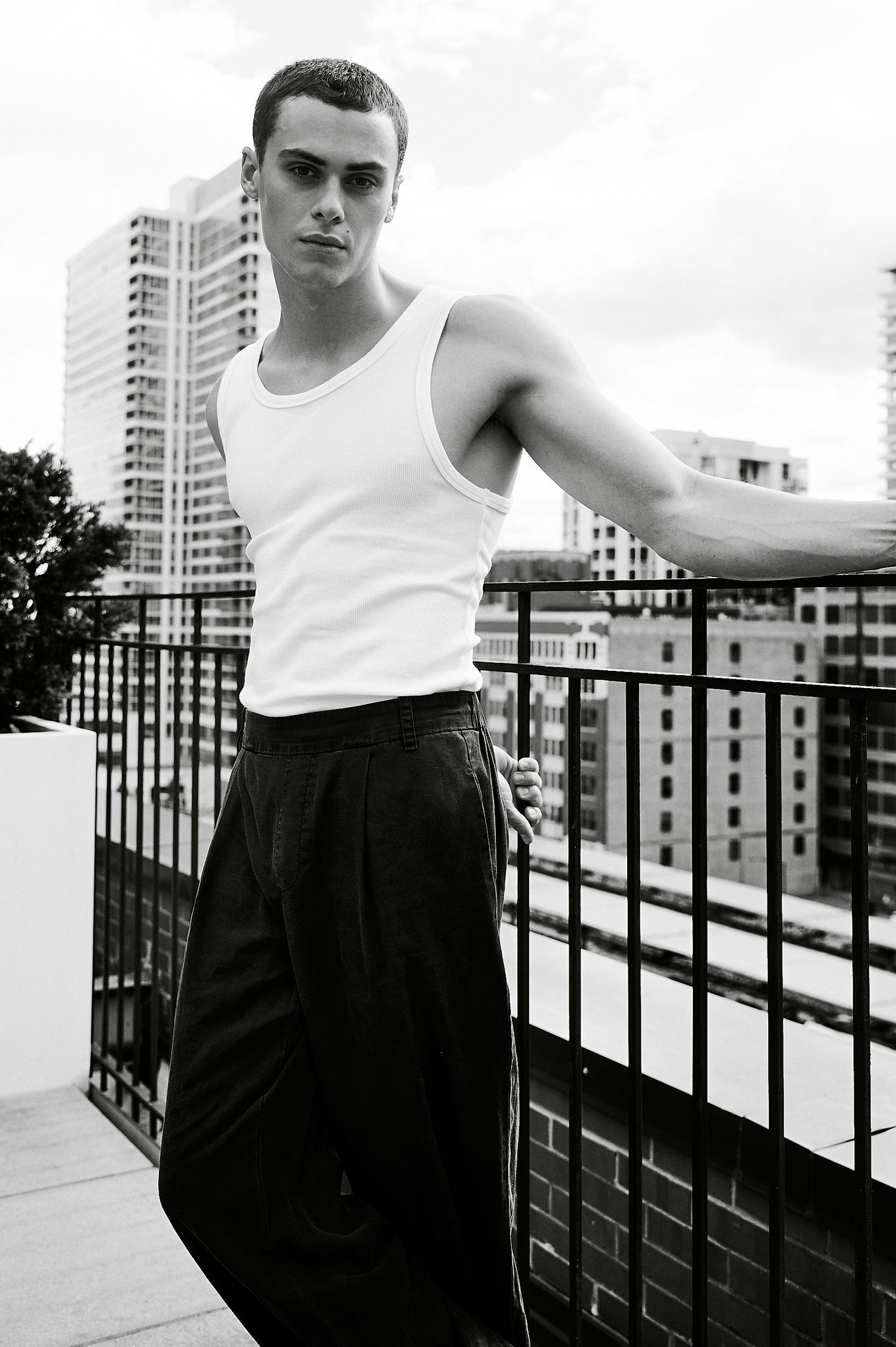 Young man in white tank top and dark trousers posing on urban rooftop balcony, black and white photo.