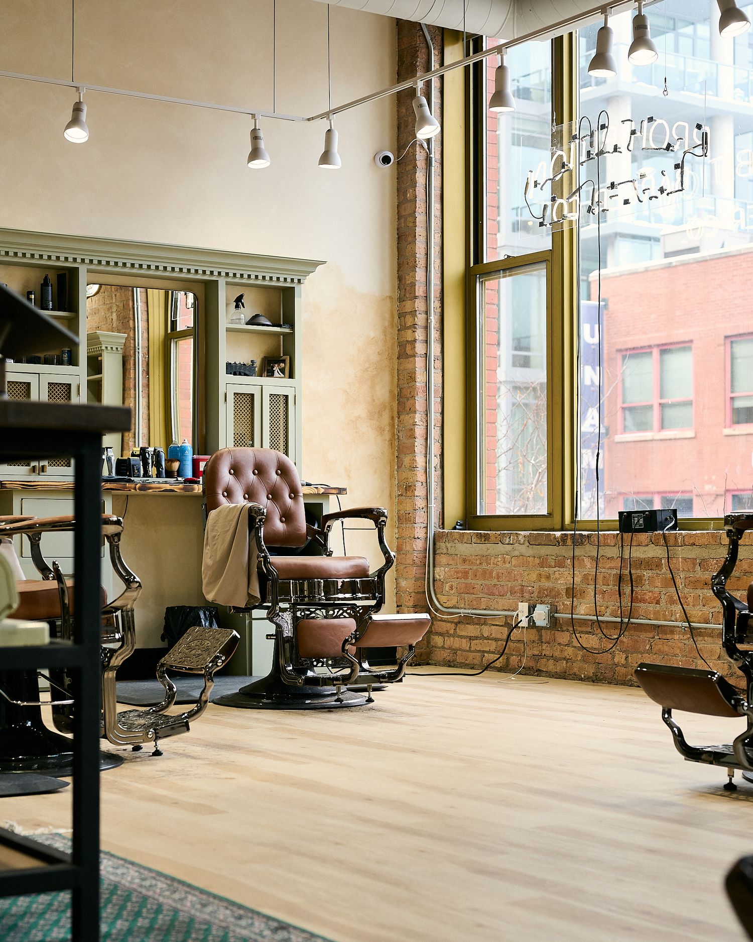 Vintage barber shop interior with leather chairs, exposed brick walls, hardwood floors, and large windows overlooking city buildings.