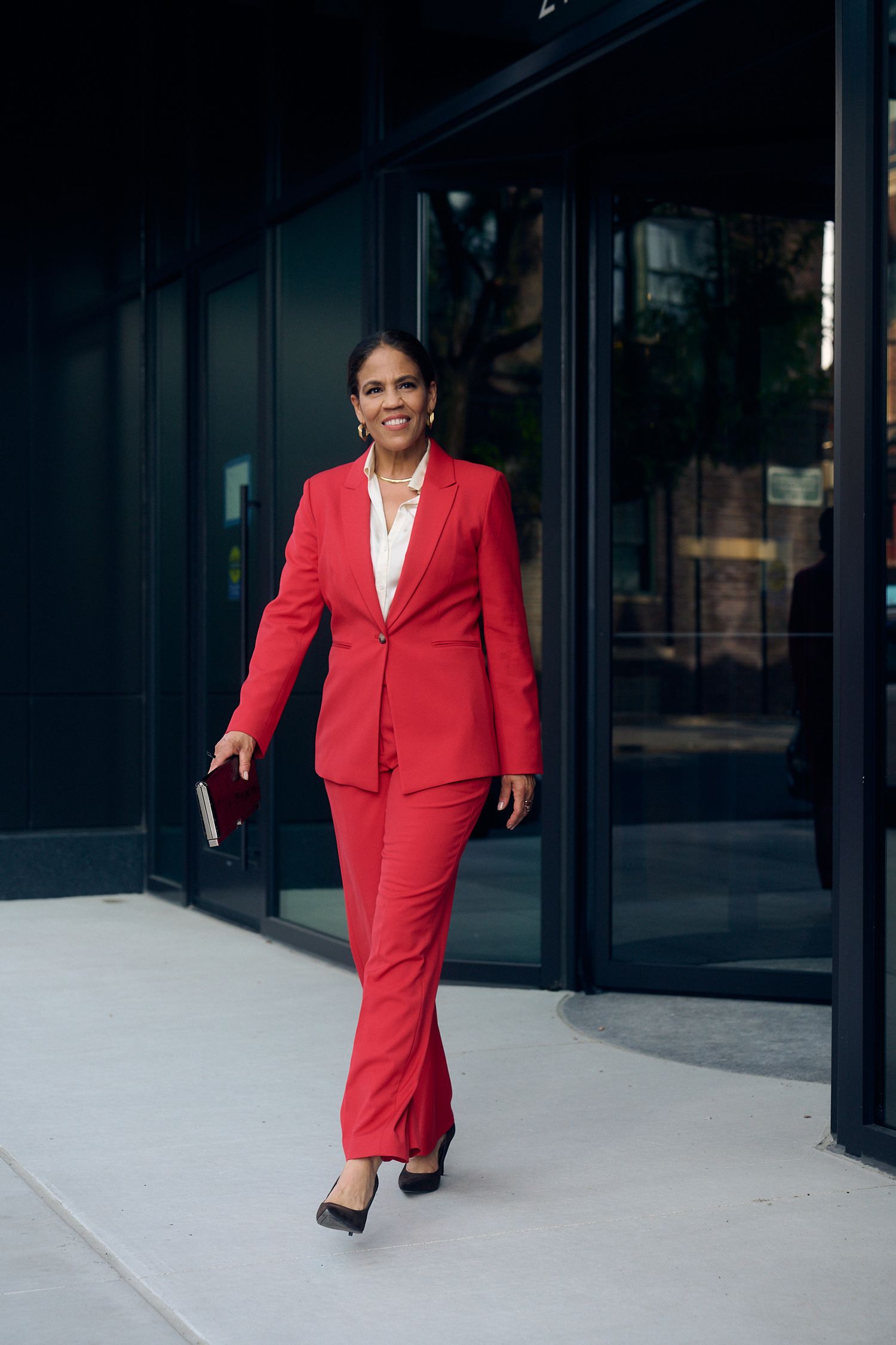 Professional woman in red pantsuit walking confidently outside modern building, holding a notebook.