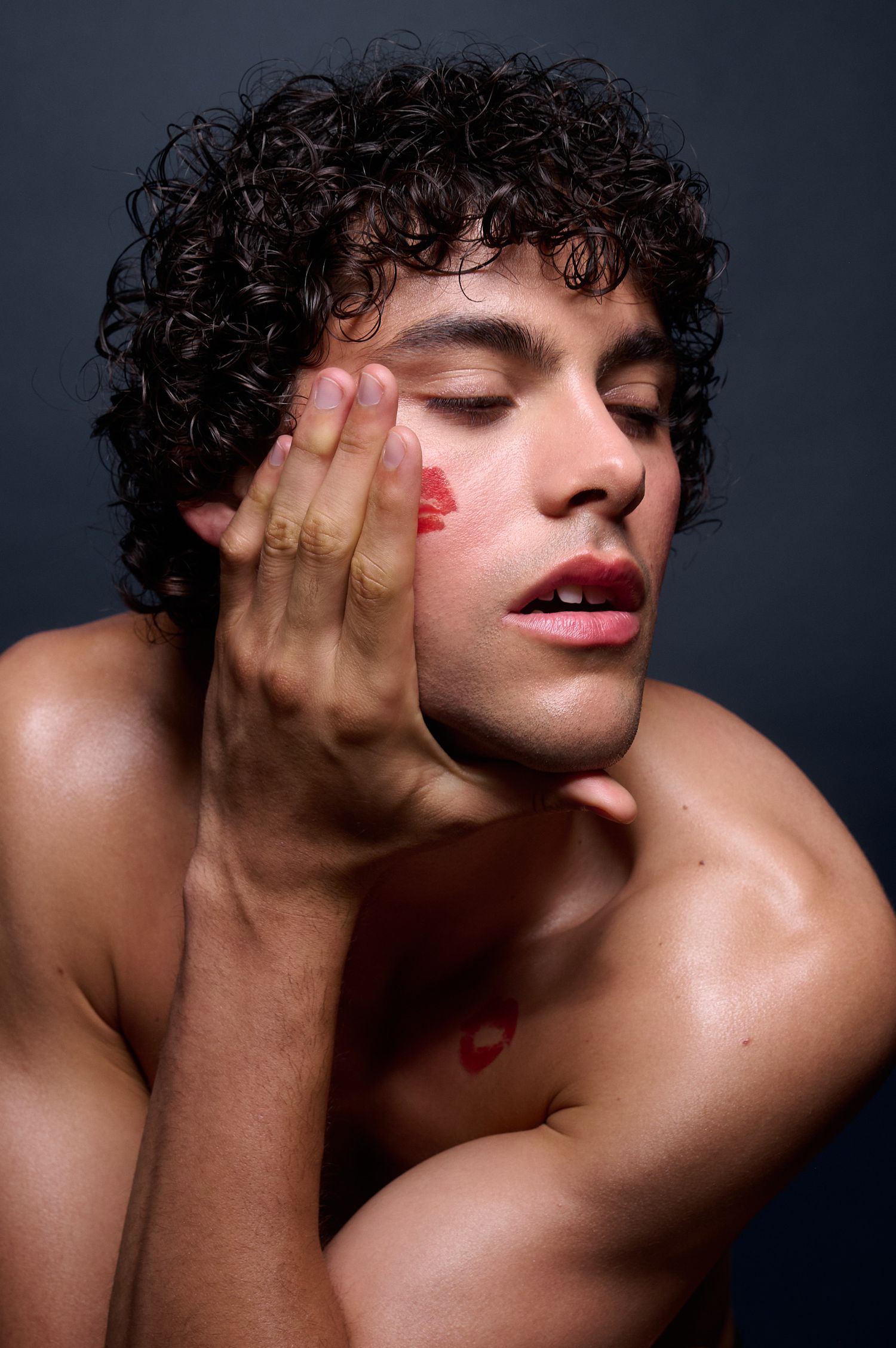 Young man with curly hair and red lipstick marks on face with eyes closed.