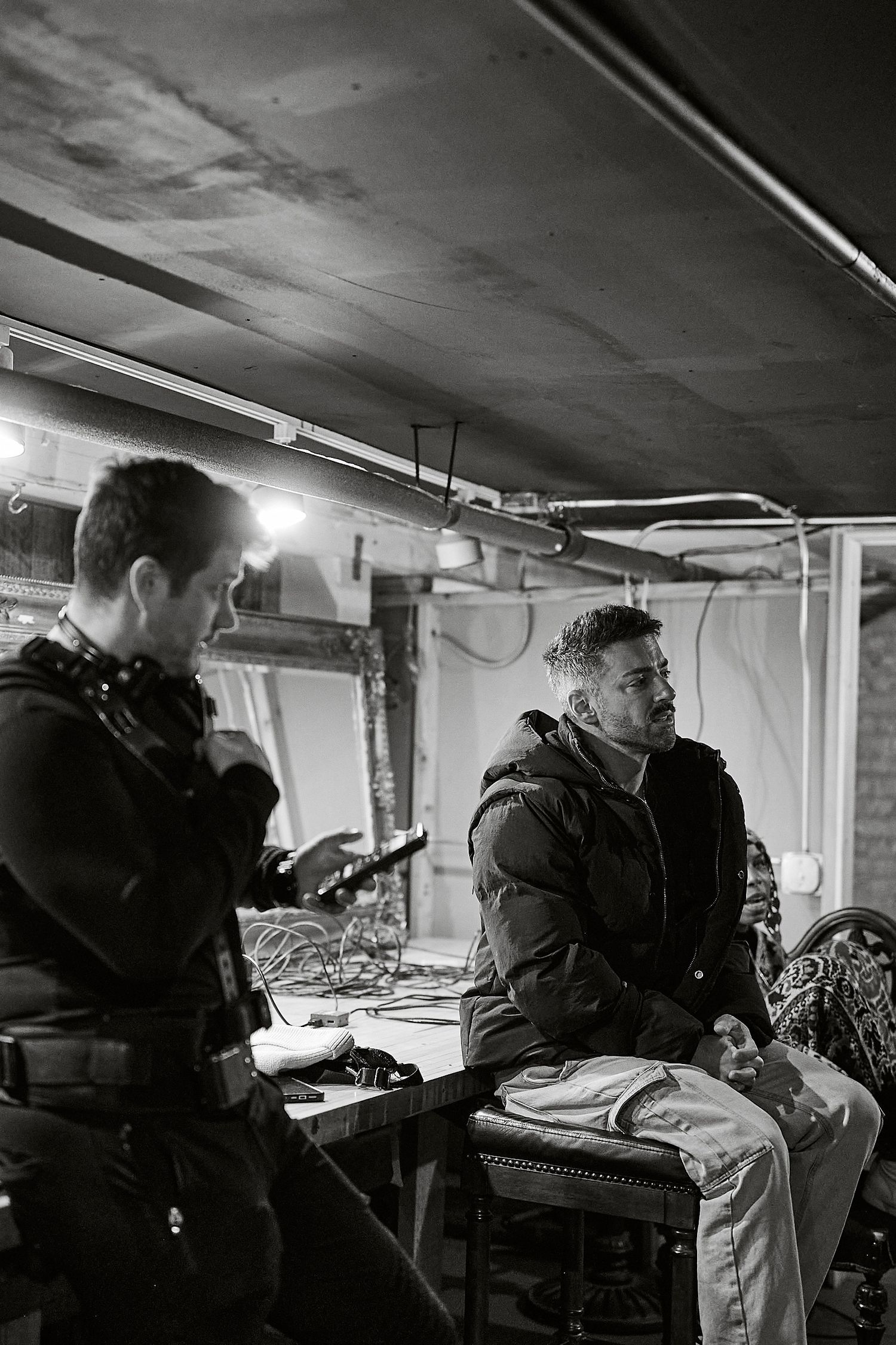 Two men in a dimly lit basement studio, one standing with a mic, the other seated at a table in a puffer jacket.