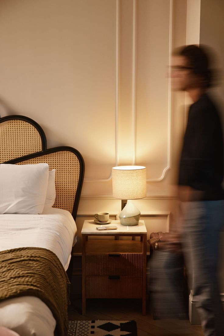 Warm bedroom with rattan headboard, wooden nightstand, glowing table lamp, and a blurred person walking past.