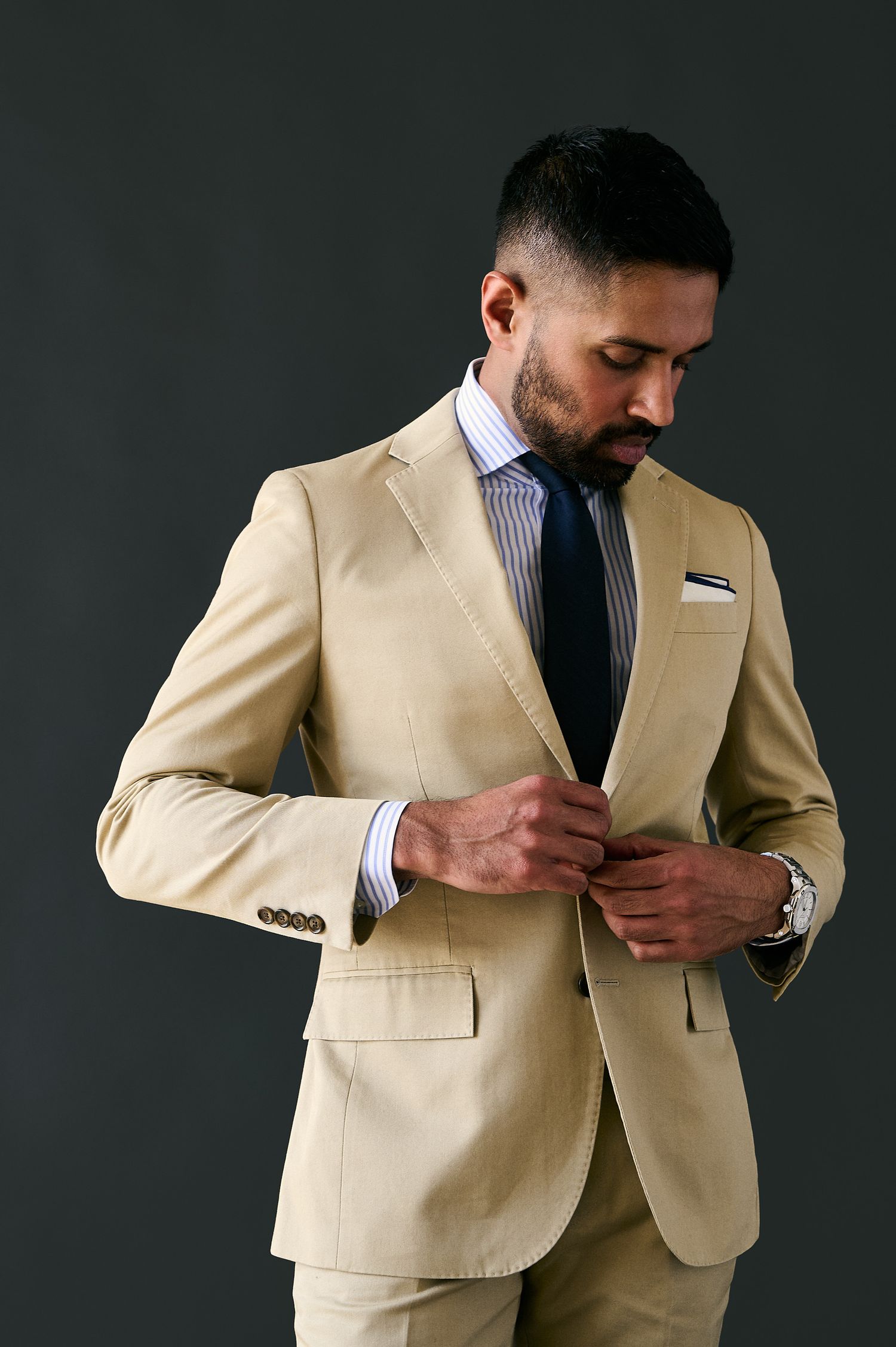 Man in beige suit adjusting jacket button, wearing navy tie and white pocket square against dark background.