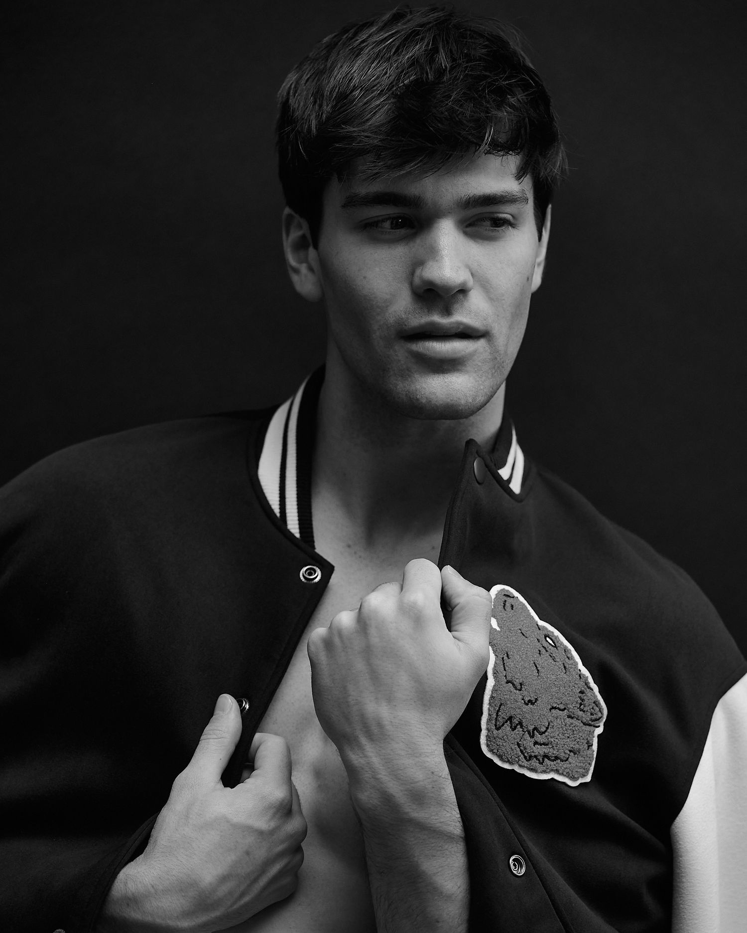 Black and white photo of a young man in a varsity jacket against a dark background.