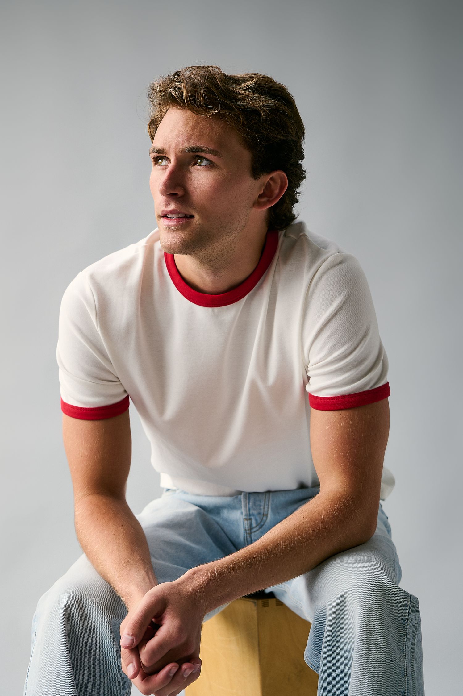 Young man in white red-trim ringer tee and light jeans sits on wooden stool, looking away thoughtfully.