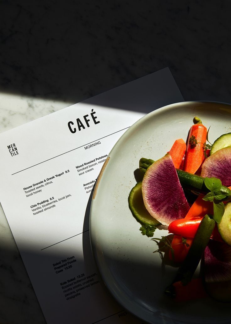 A colorful vegetable salad on a white plate beside a cafe menu on a marble surface in natural light.