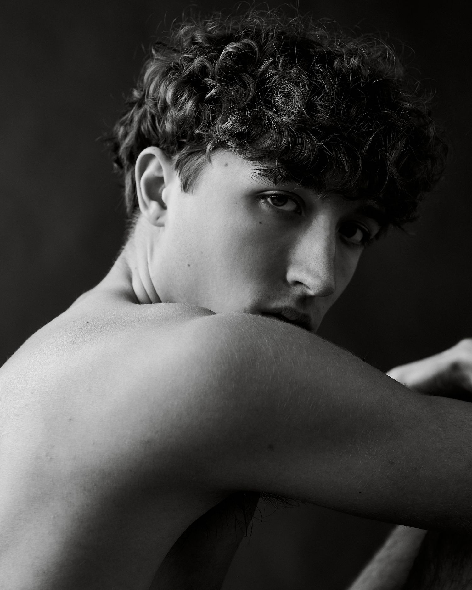 Black and white portrait of a young man with curly hair resting his chin on his arm, gazing at camera.