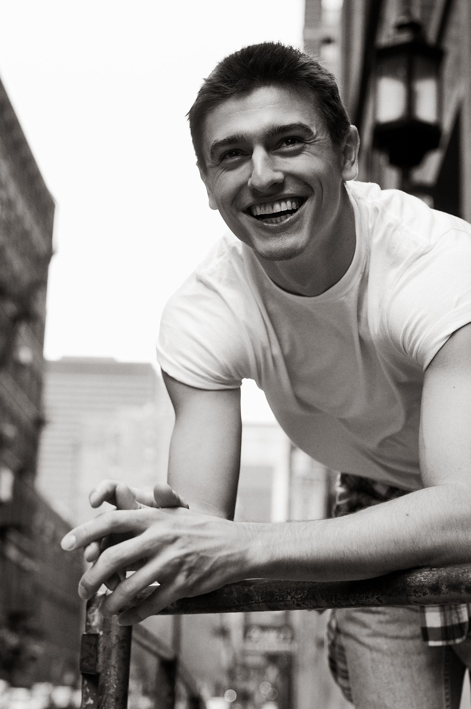 Young man in white t-shirt leaning on railing outdoors, smiling broadly in black and white urban photo.