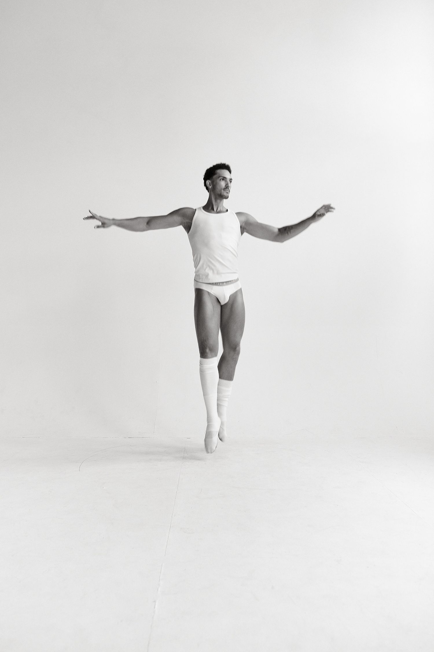 Male ballet dancer in white tank and briefs performing an arabesque with arms extended in a bright studio.