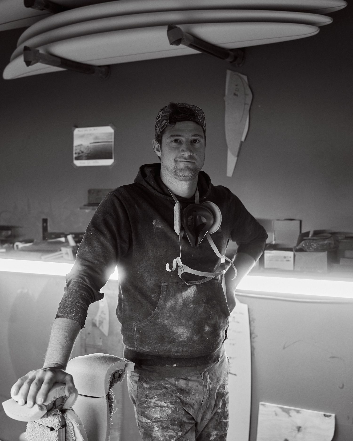 A surfboard shaper poses confidently in his dusty workshop with boards mounted on the wall.