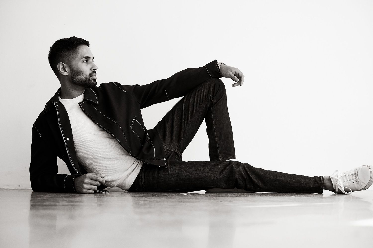 Black and white photo of a stylish man sitting on floor in dark jeans and jacket, posing confidently.
