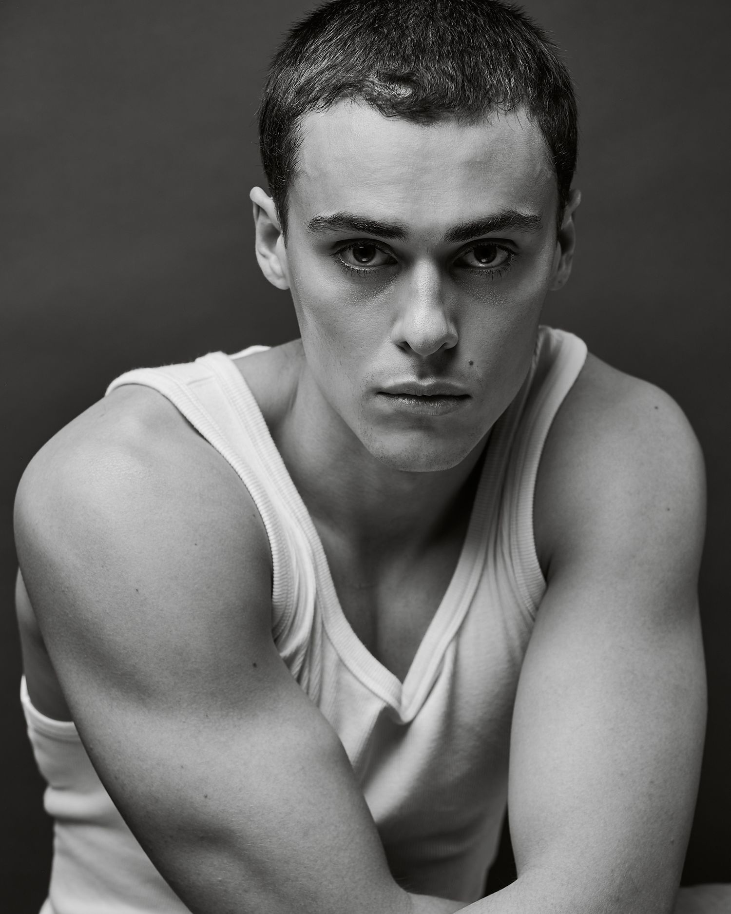 Black and white portrait of a young man in a white tank top with short hair, posing with arms crossed.
