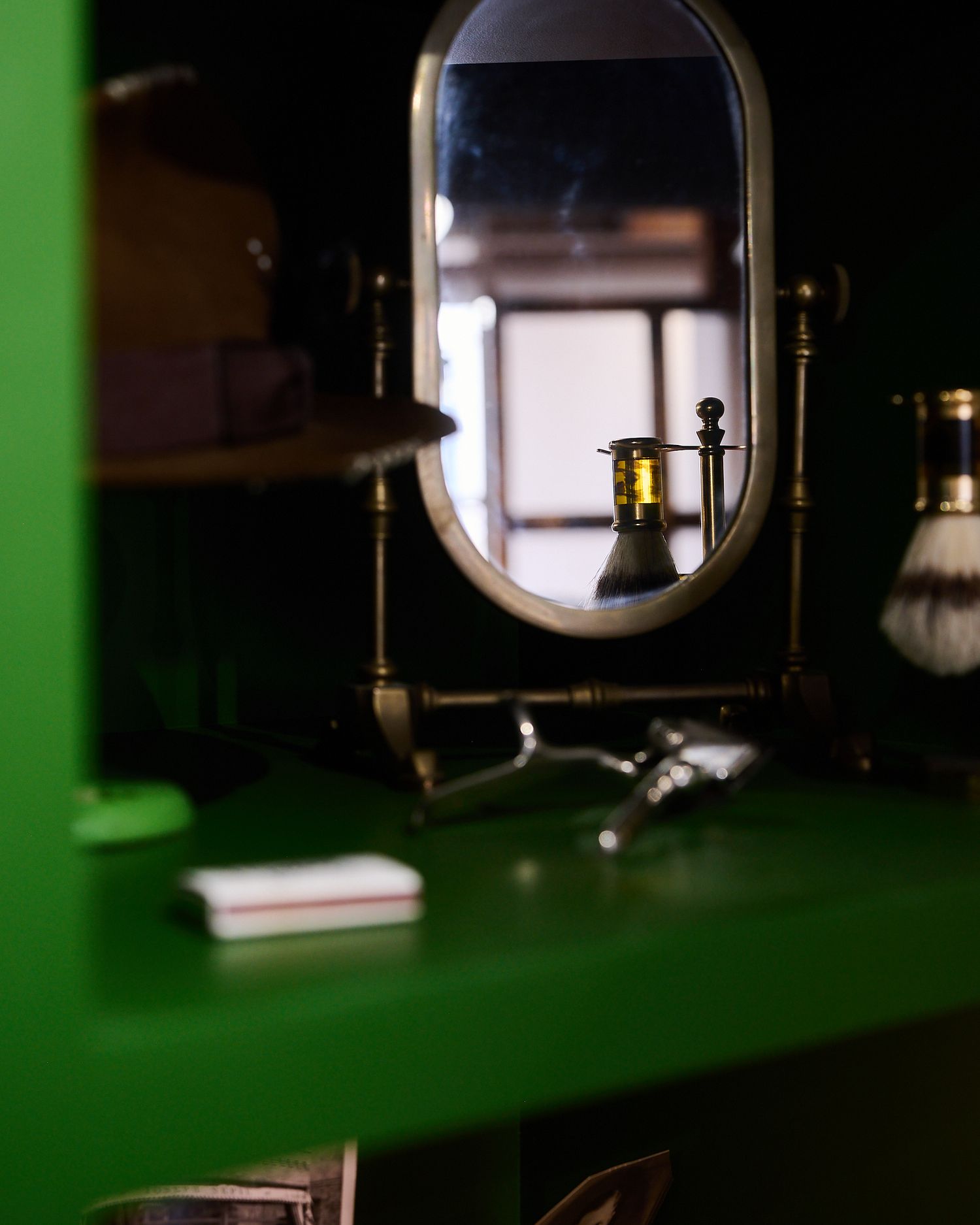 Vintage brass barber mirror on a green shelf with shaving brush and razor grooming tools in a classic barbershop.