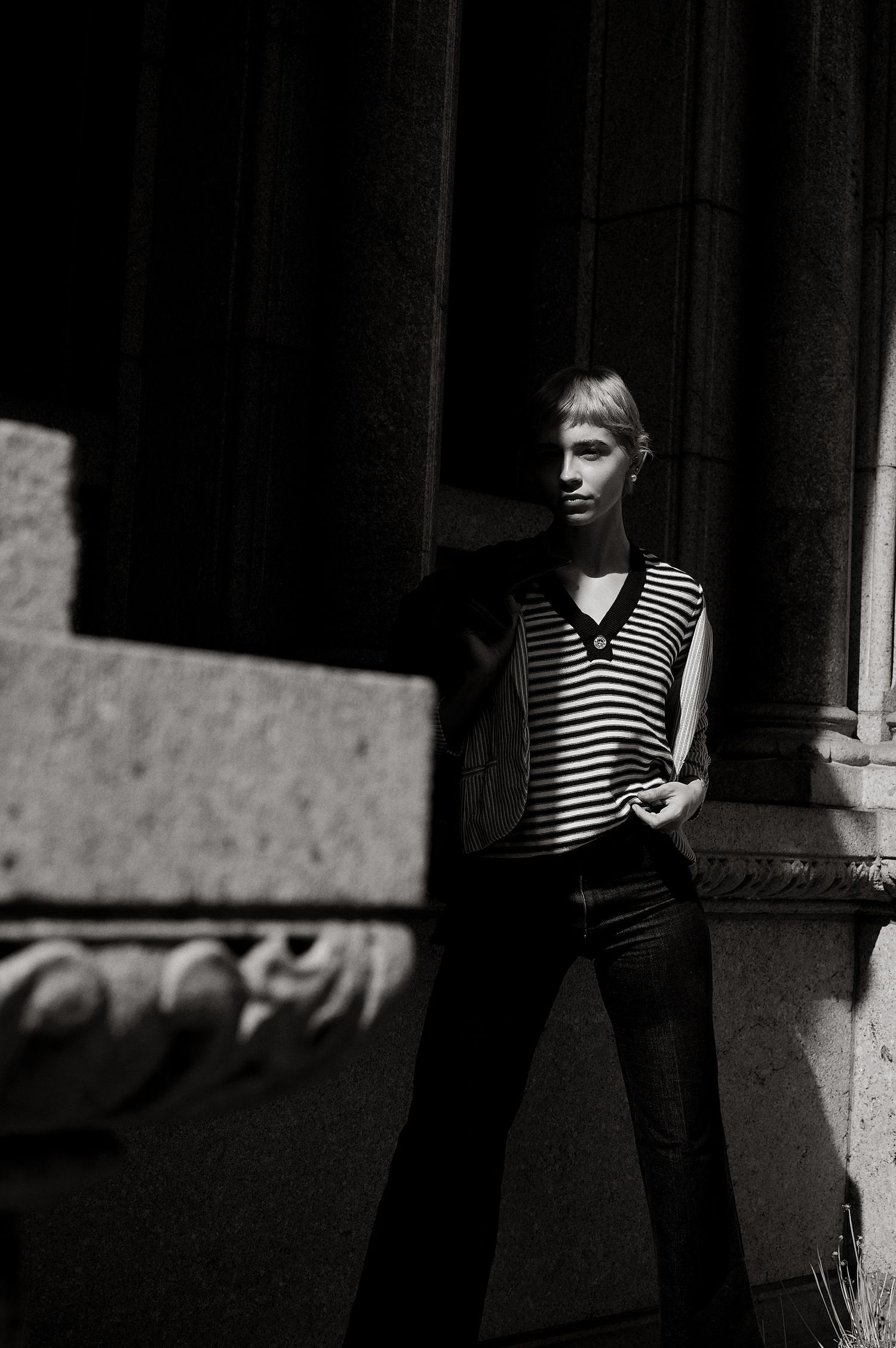 Black and white portrait of a blonde woman in striped sleeveless top and dark jeans leaning against stone columns.