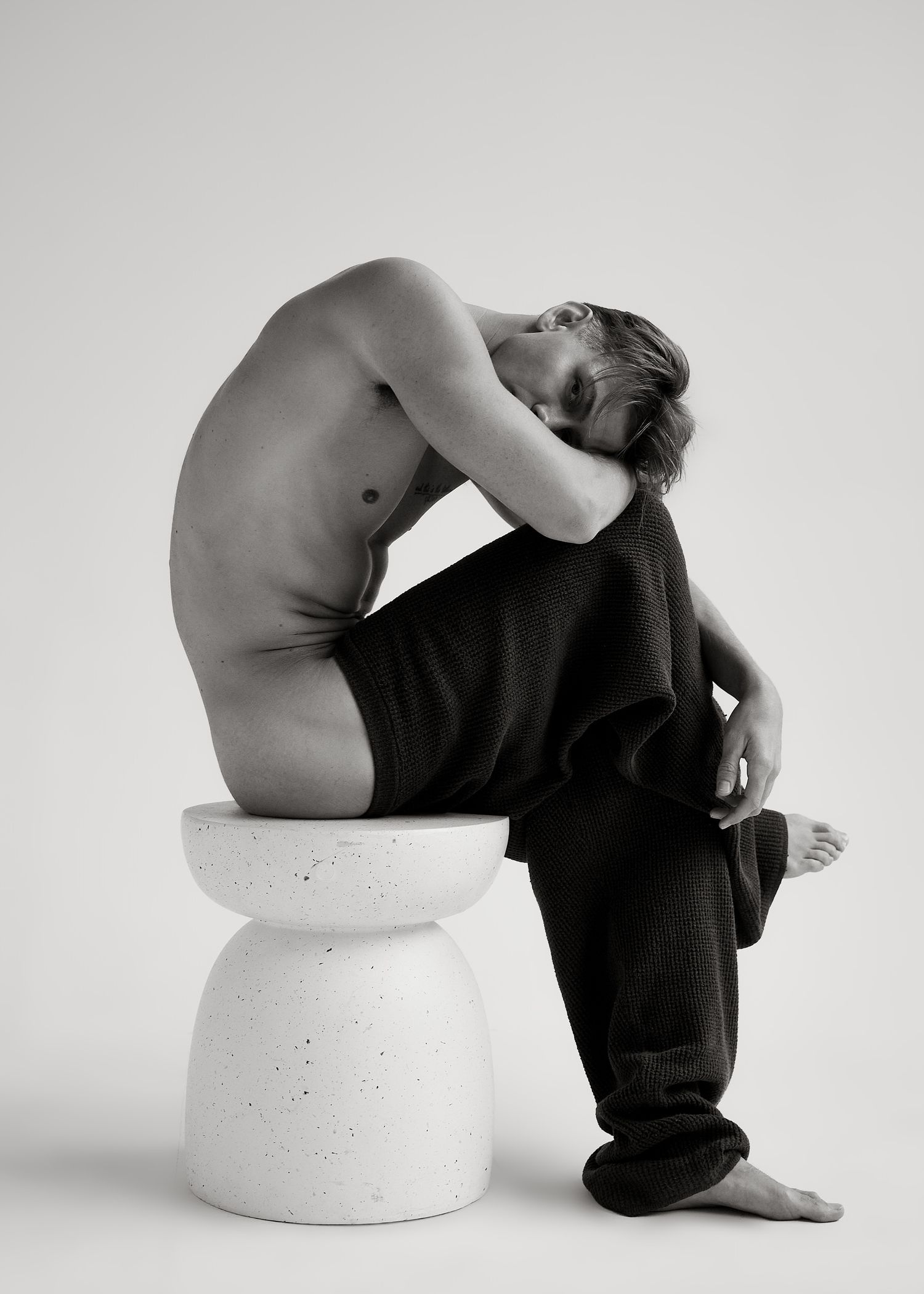 Black and white photo of a shirtless man in dark pants, resting on a white pedestal stool in a contemplative pose.