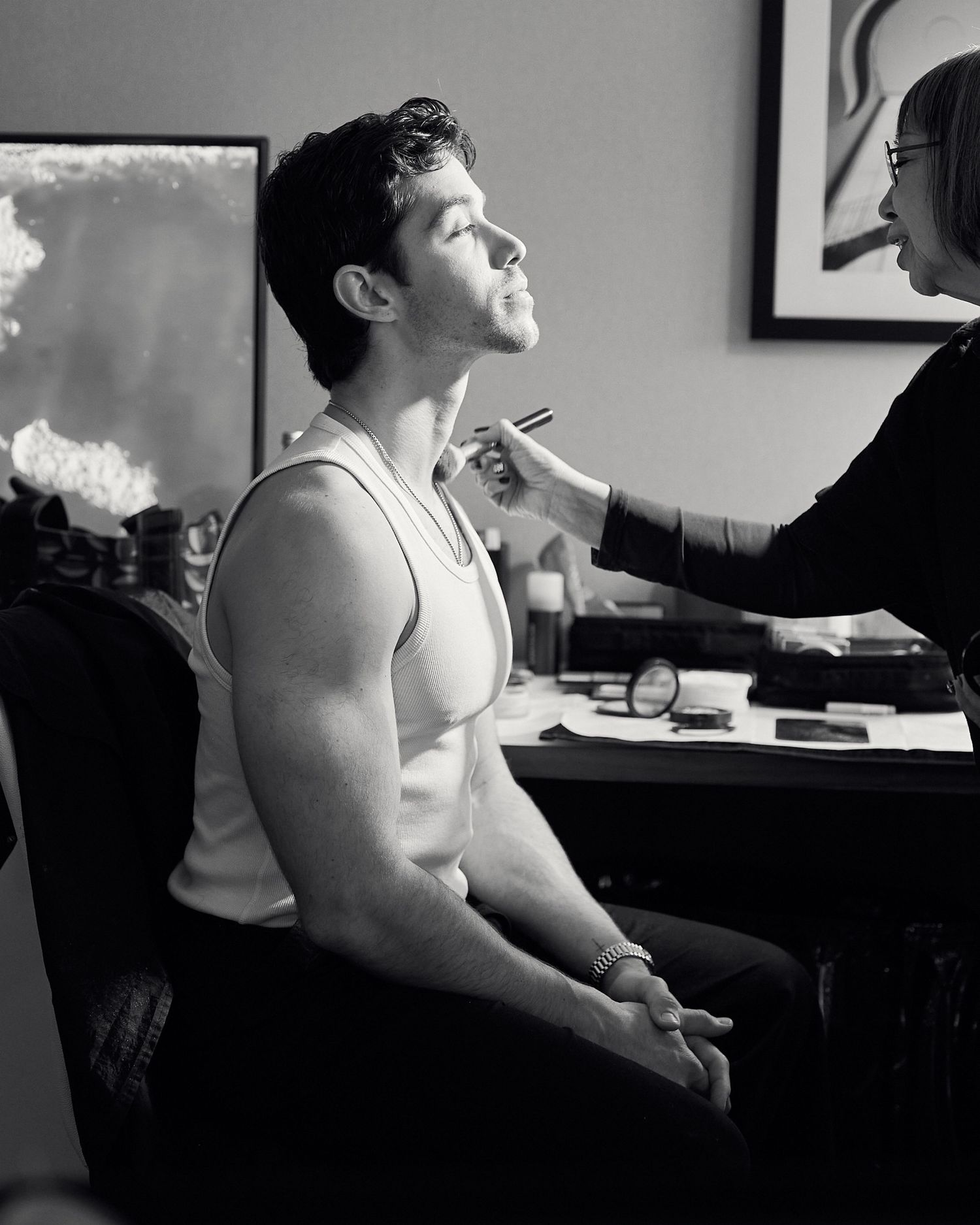 Black and white photo of a muscular man in a tank top getting makeup applied by a makeup artist.