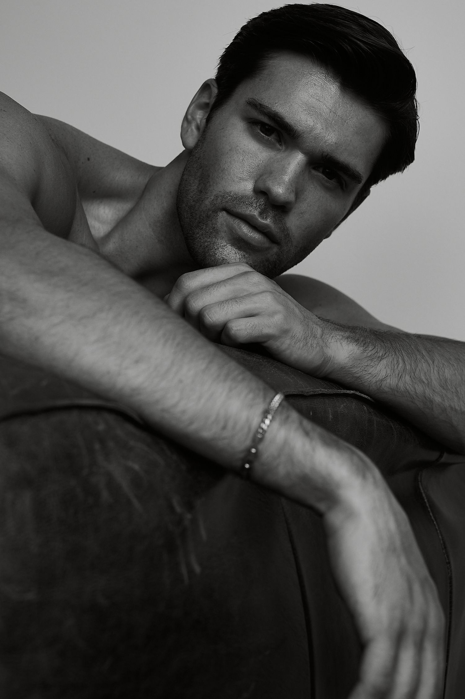 Black and white portrait of a shirtless man resting his arms on a sofa, wearing a bracelet, with a serious expression.