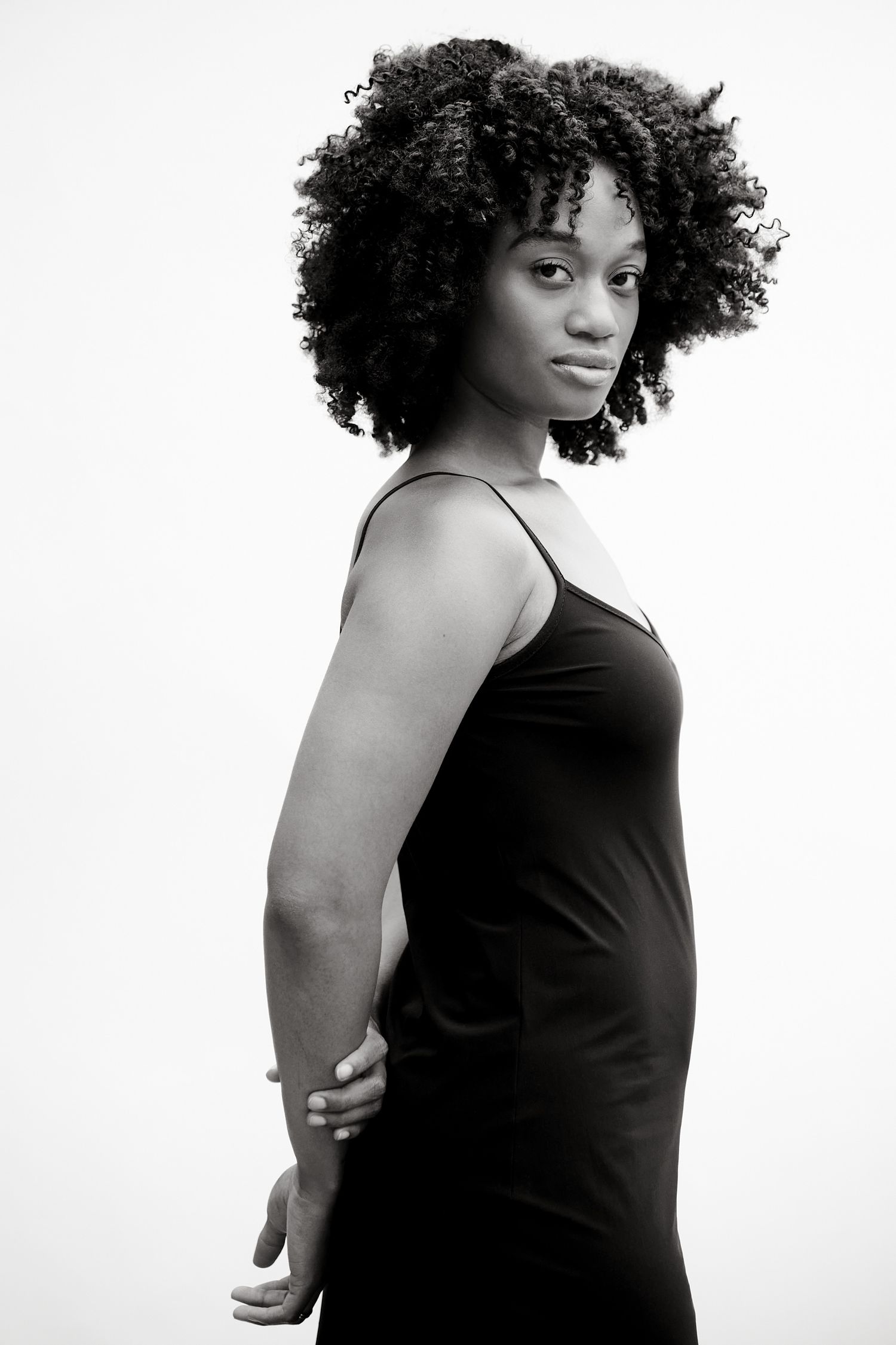 Black and white portrait of a young woman with natural afro hair wearing a black slip dress, posing against white background.