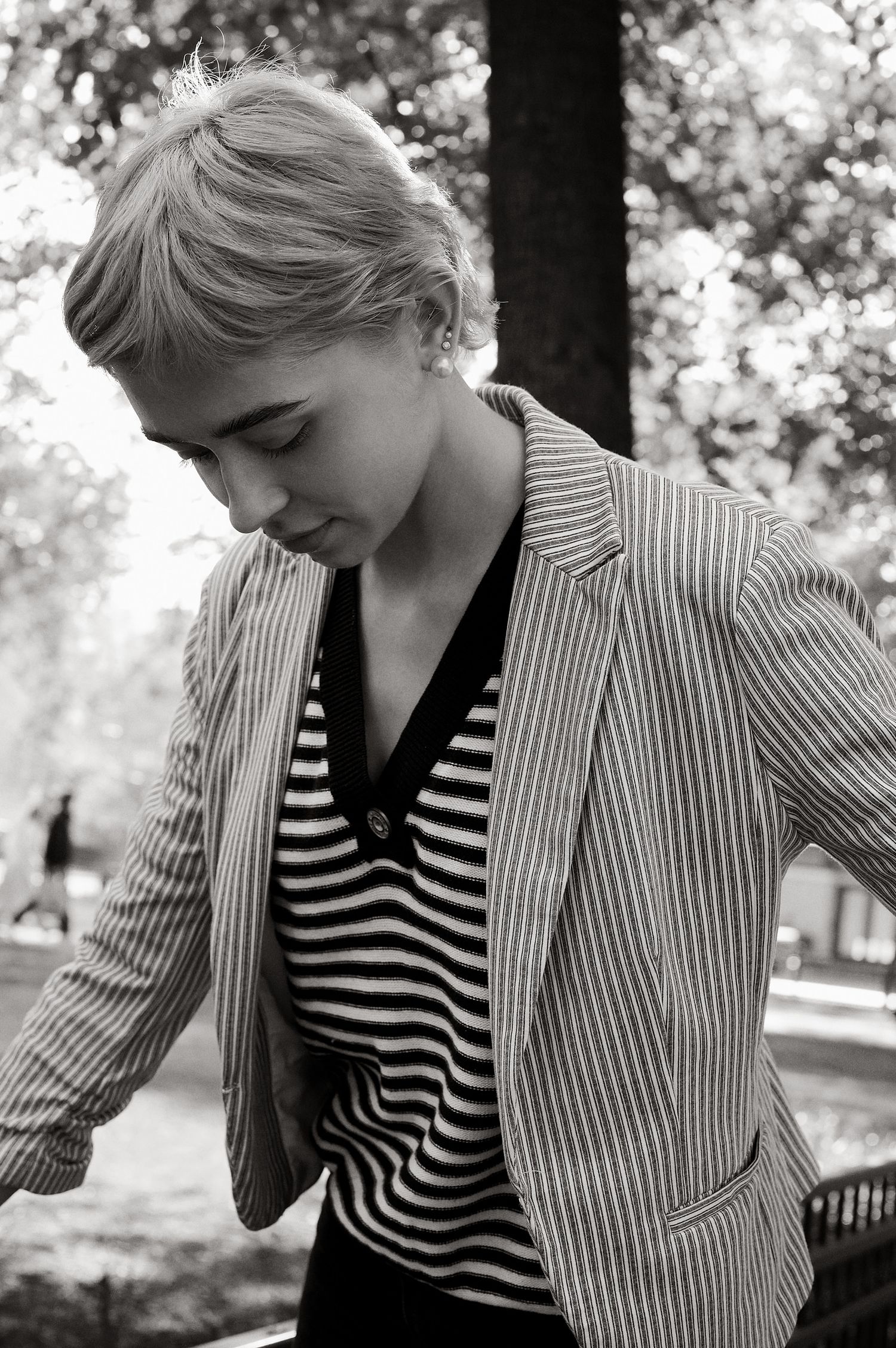 Young blonde woman in striped blazer and striped top looks down thoughtfully in black and white outdoor portrait.