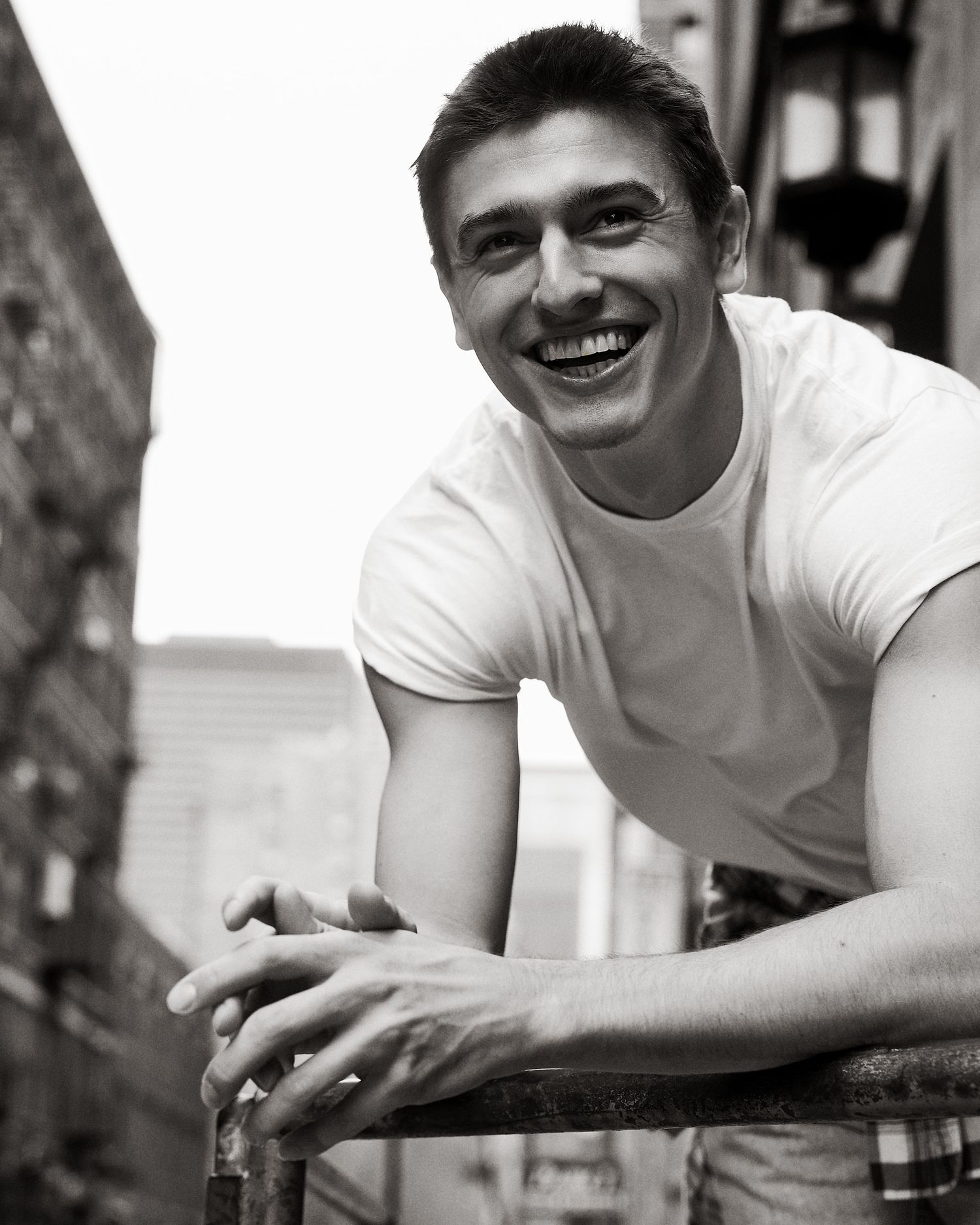 Smiling young man in white t-shirt leaning on railing in black and white urban photo.