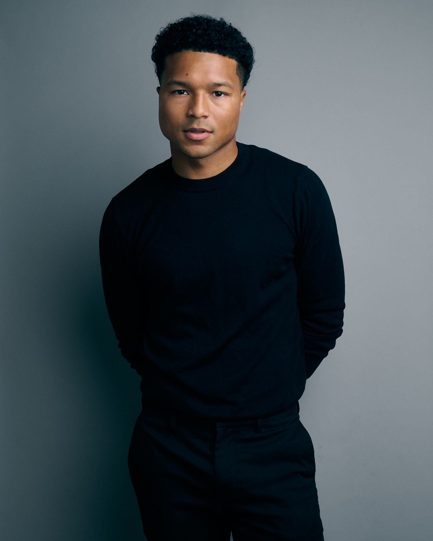 Full body portrait of a young Black man in black sweater and trousers against a gray background.