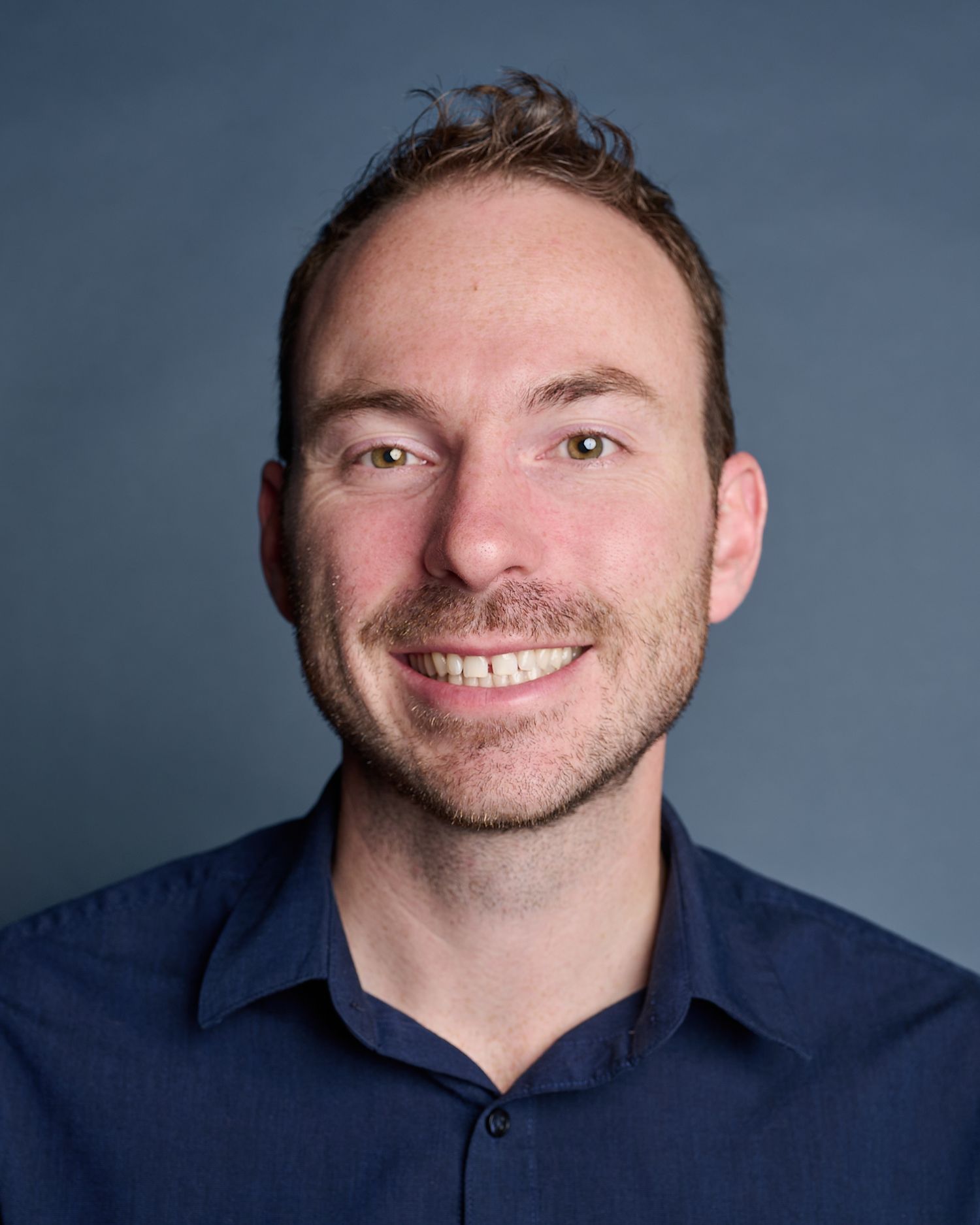 Smiling man with short brown hair and green eyes wearing a navy blue shirt against a gray background.
