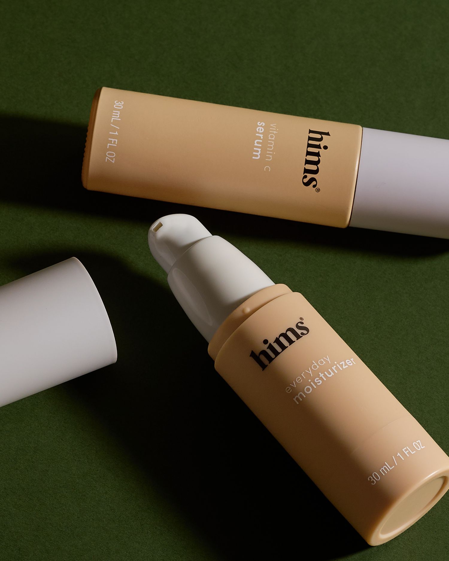 Two Hims skincare products, a serum and daily moisturizer, displayed on a dark green surface.