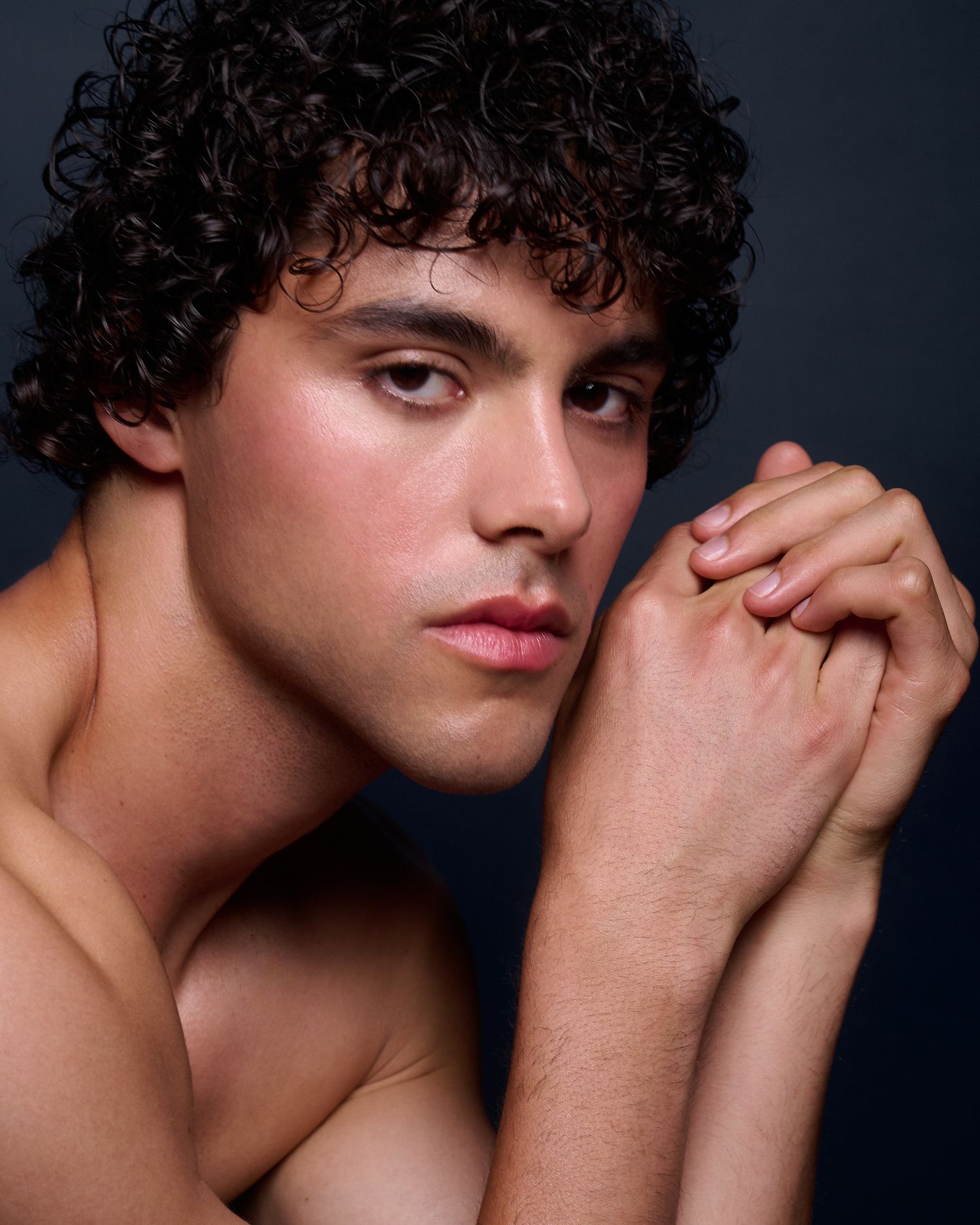 Young man with curly dark hair poses shirtless with hands clasped near face against dark background.