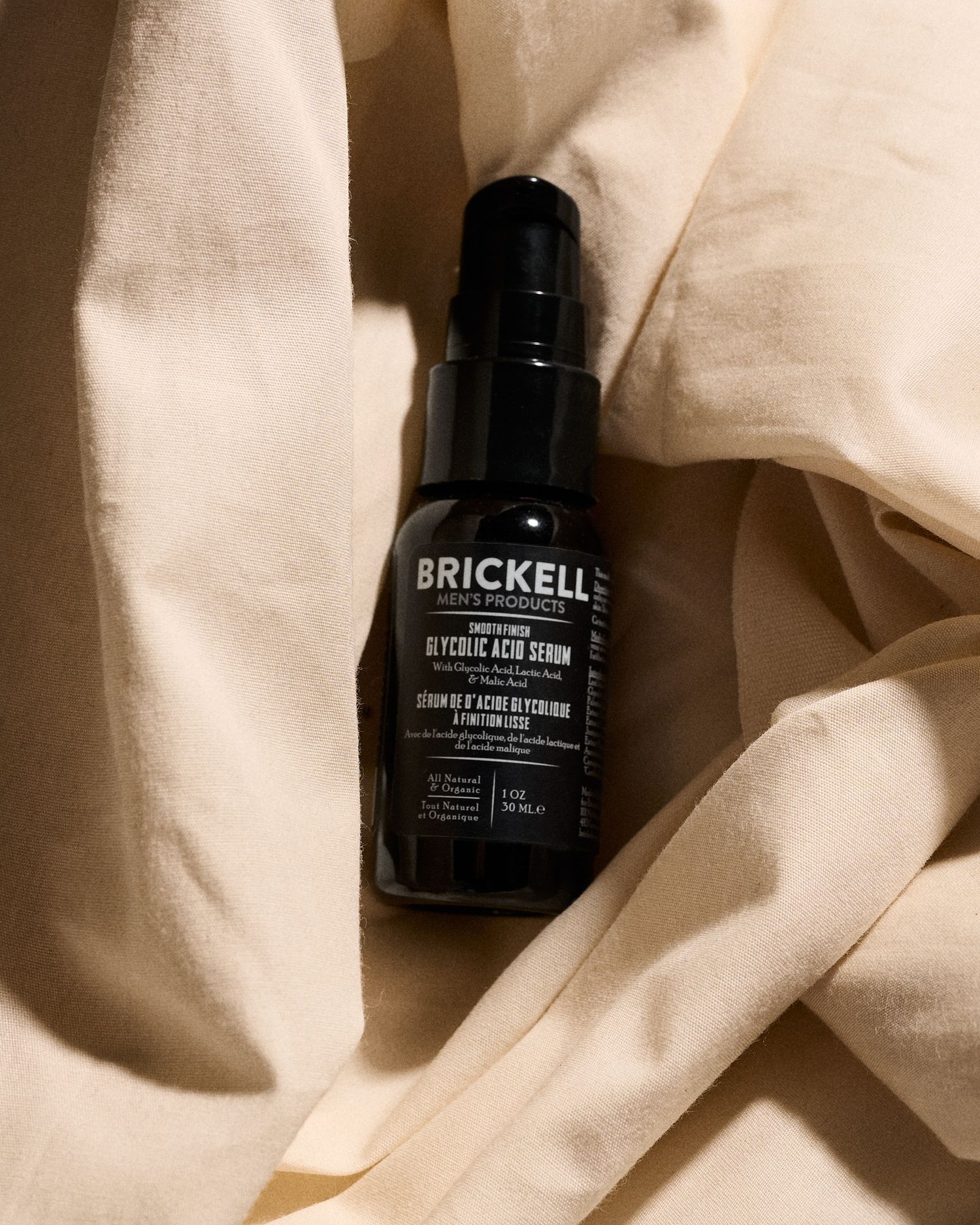 Brickell Men's Glycolic Acid Serum in dark glass bottle displayed on cream linen fabric.