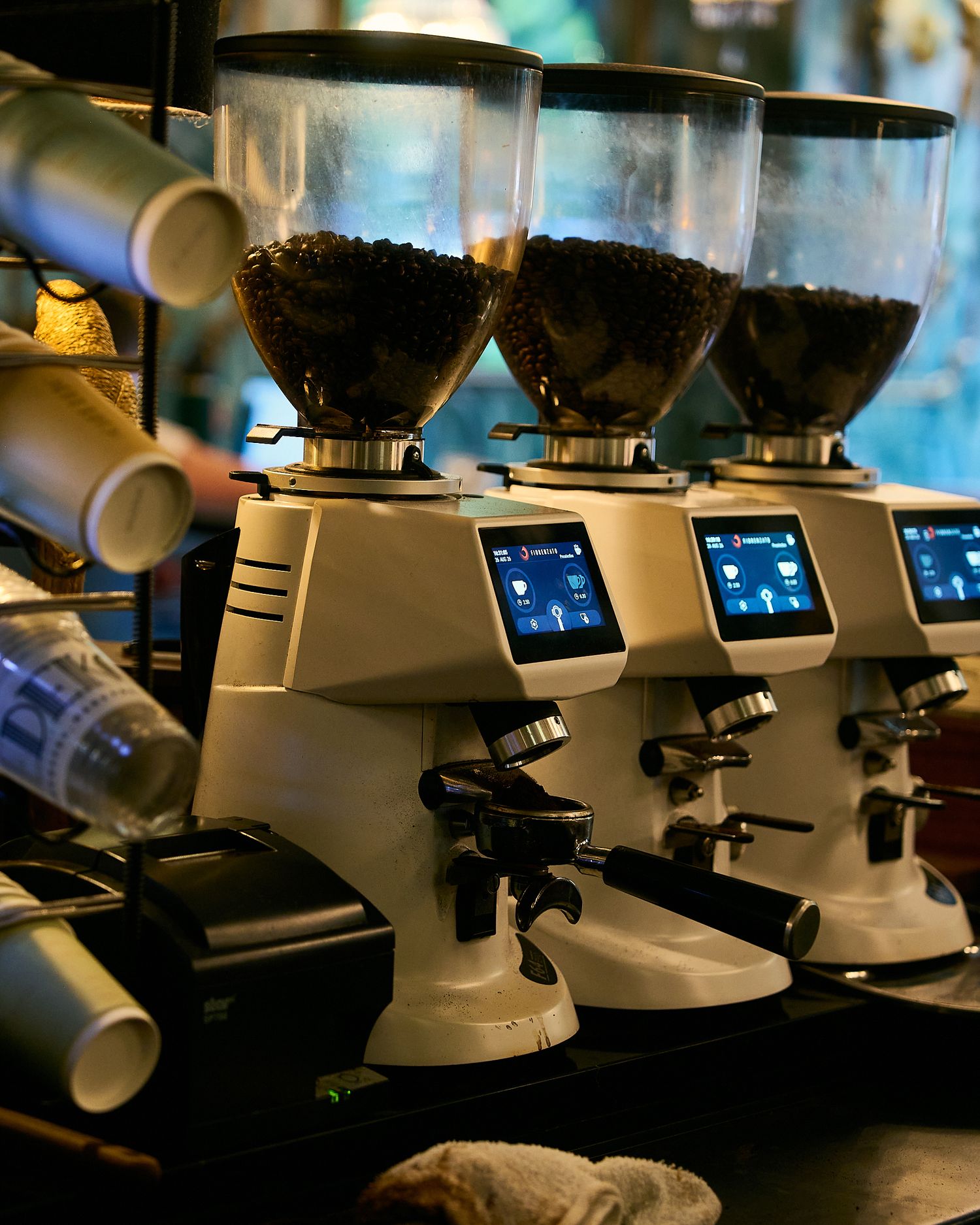 Three commercial coffee grinders with digital displays filled with whole beans on a cafe counter.